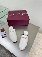 White mule shoes with gold horsebit hardware, pair displayed beside Gucci box