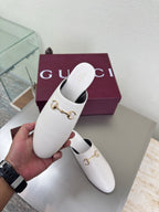 White mule shoe with gold horsebit detail, held in hand with matching pair