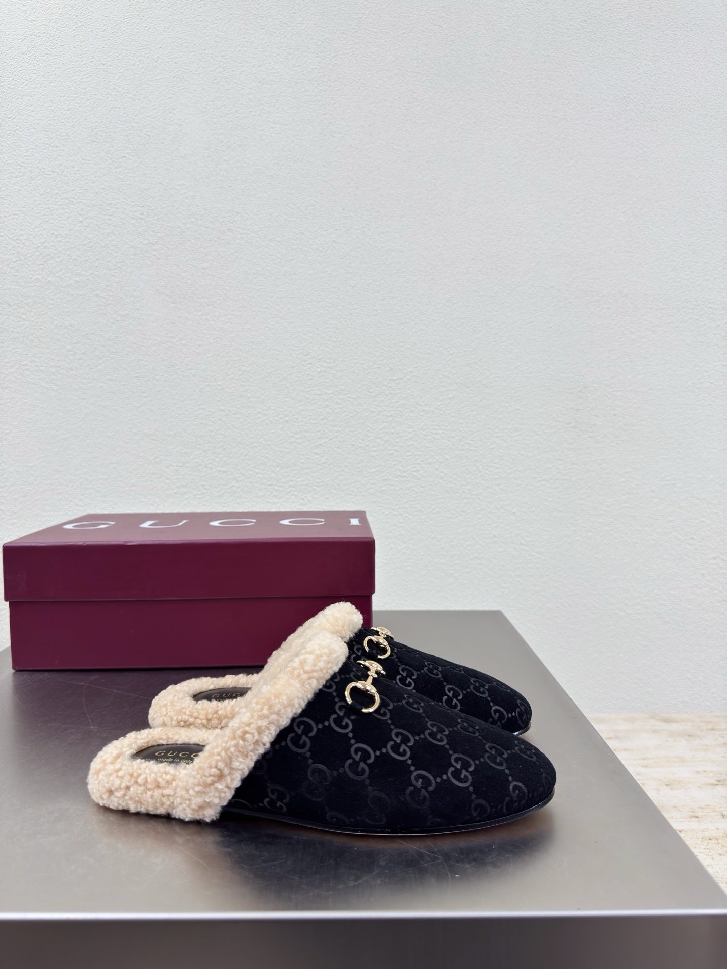 Black Gucci mule with beige shearling lining and GG pattern, beside maroon Gucci box.