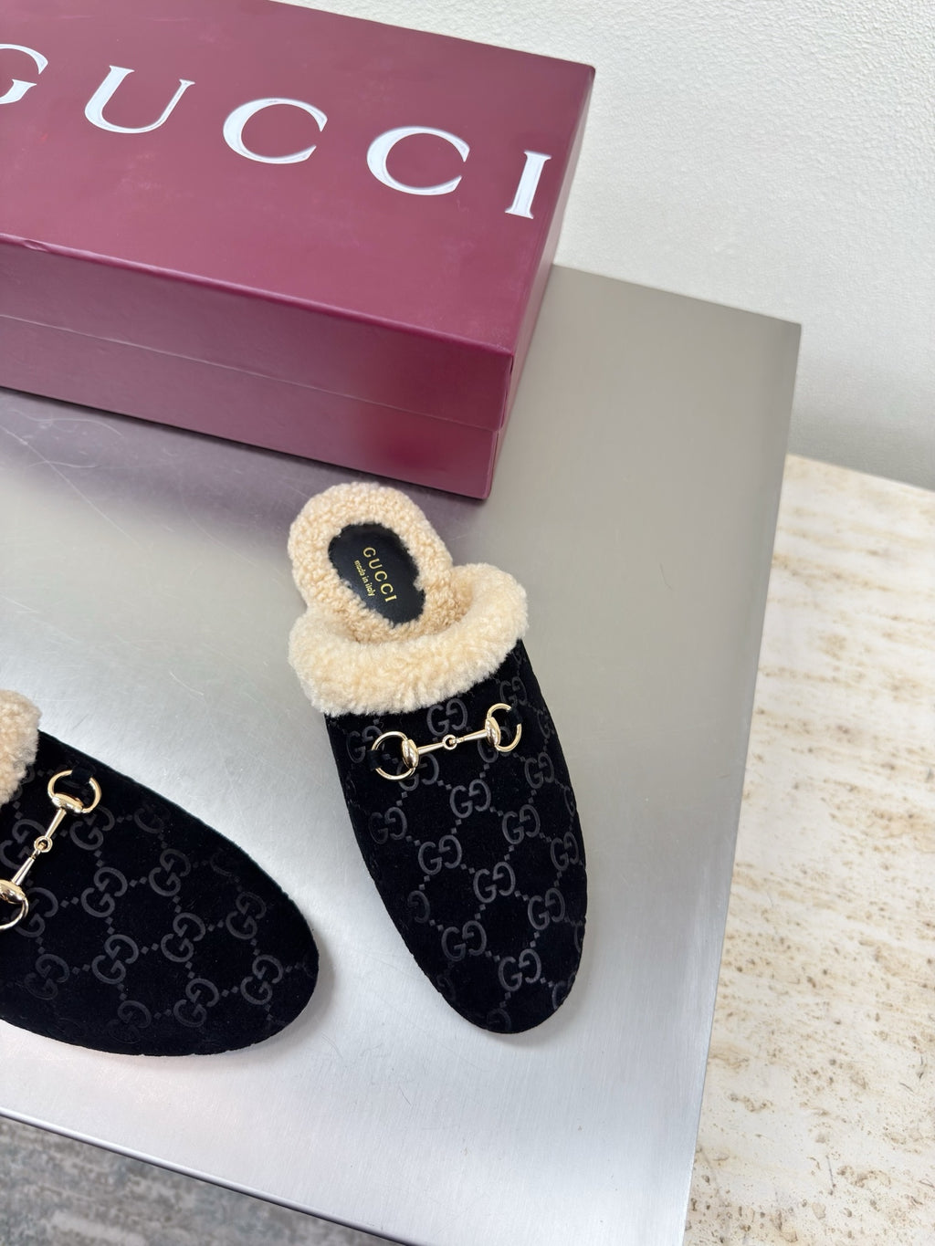 Black Gucci mules with beige shearling lining and GG pattern, side view on display.