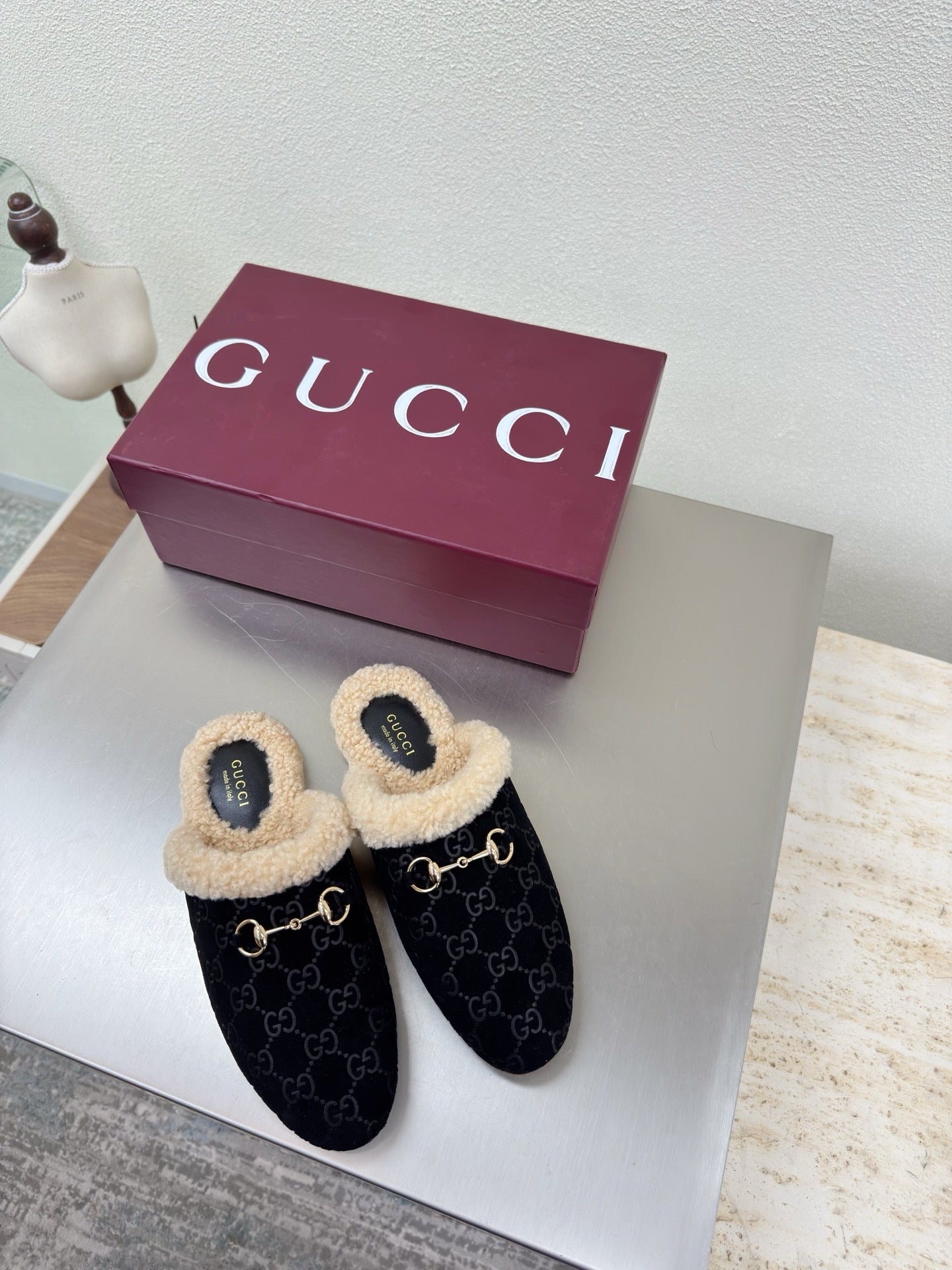 Black Gucci mules with beige shearling lining and GG pattern, on white display table.