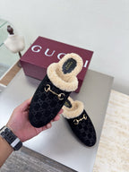 Handheld black Gucci mule with beige shearling lining and GG pattern, with maroon box behind.