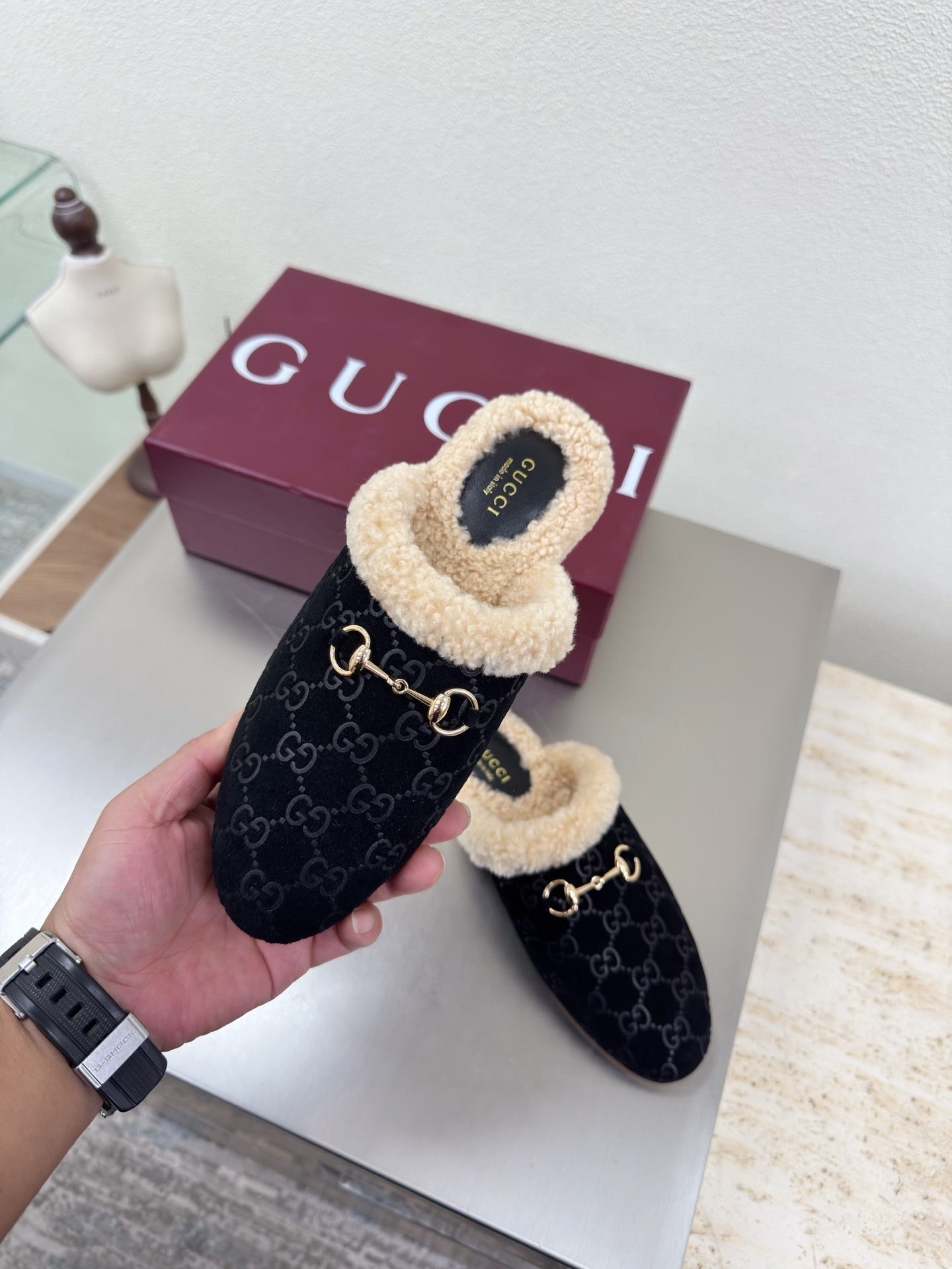 Handheld black Gucci mule with beige shearling lining and GG pattern, with maroon box behind.