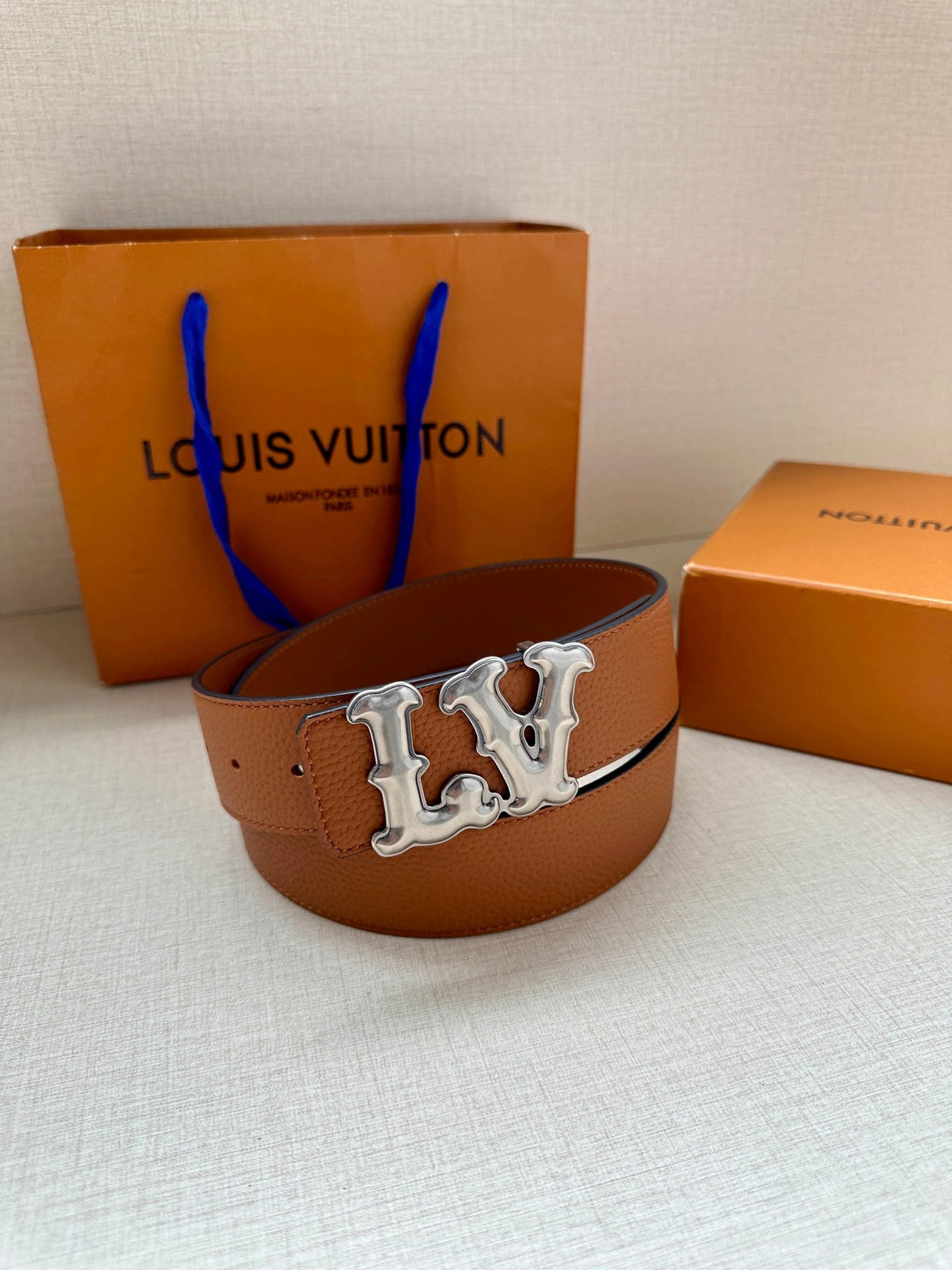 Tan leather belt with silver-toned LV buckle, belt looped on the bed