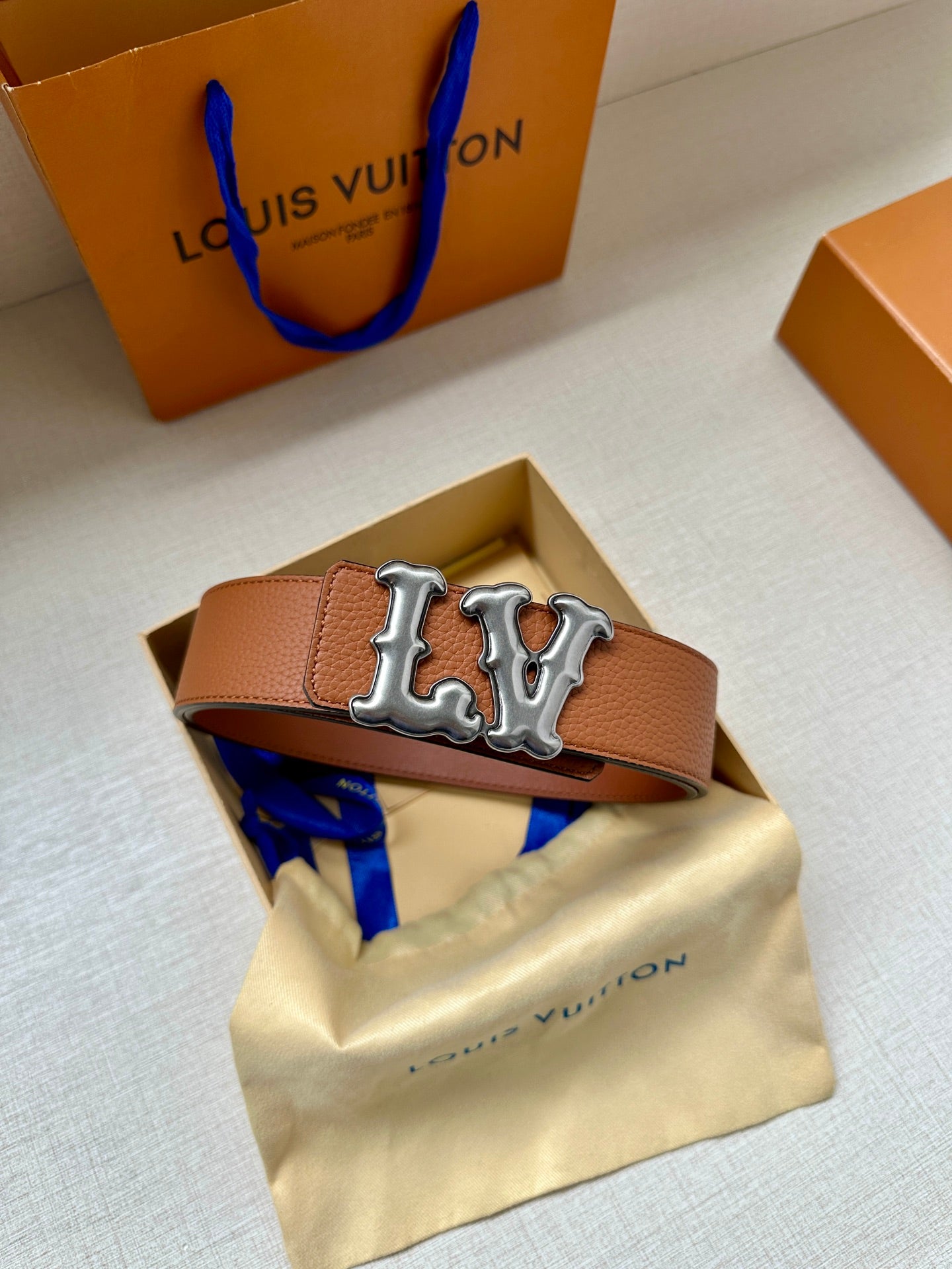 Tan leather belt with silver-toned LV buckle on top of a yellow dust bag