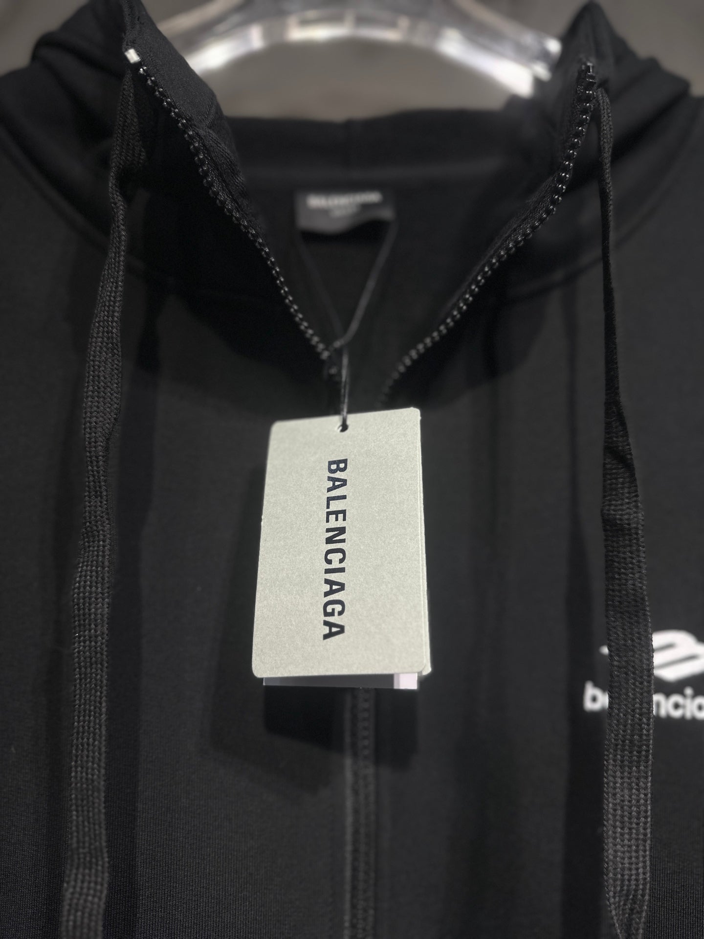 Black Balenciaga hoodie with zip front and hood, hanging with brand tag.