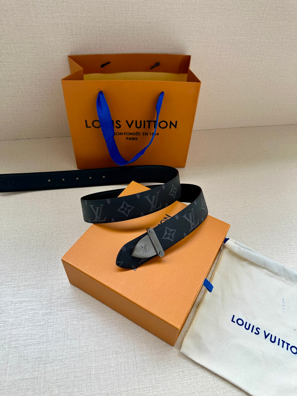 Monogram belt with dark strap, gold-tone clasp buckle, and blue tag accent