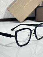 Eyeglasses close-up with black frame and gold-toned logo detail, clear lenses