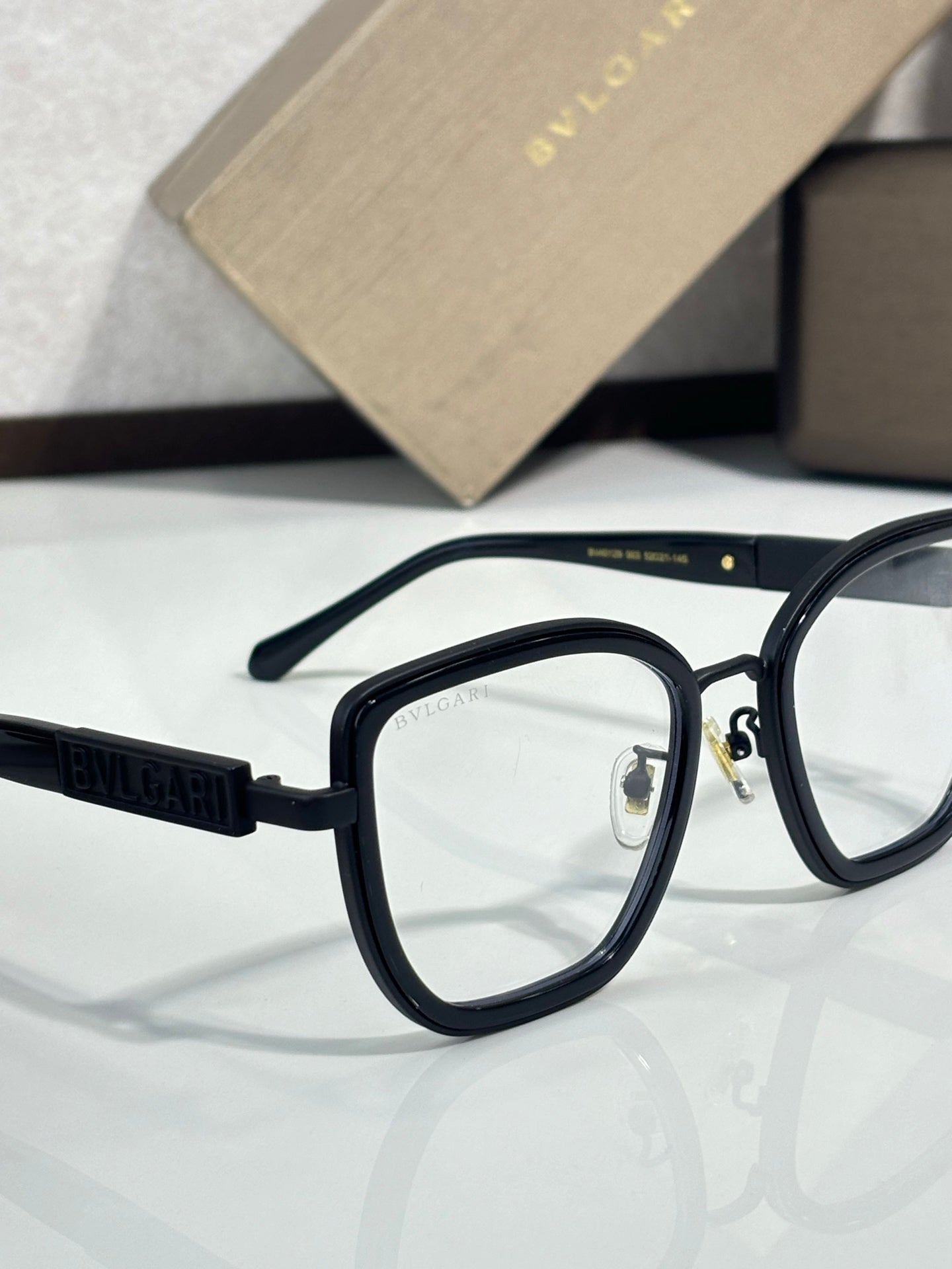 Eyeglasses close-up with black frame and gold-toned logo detail, clear lenses