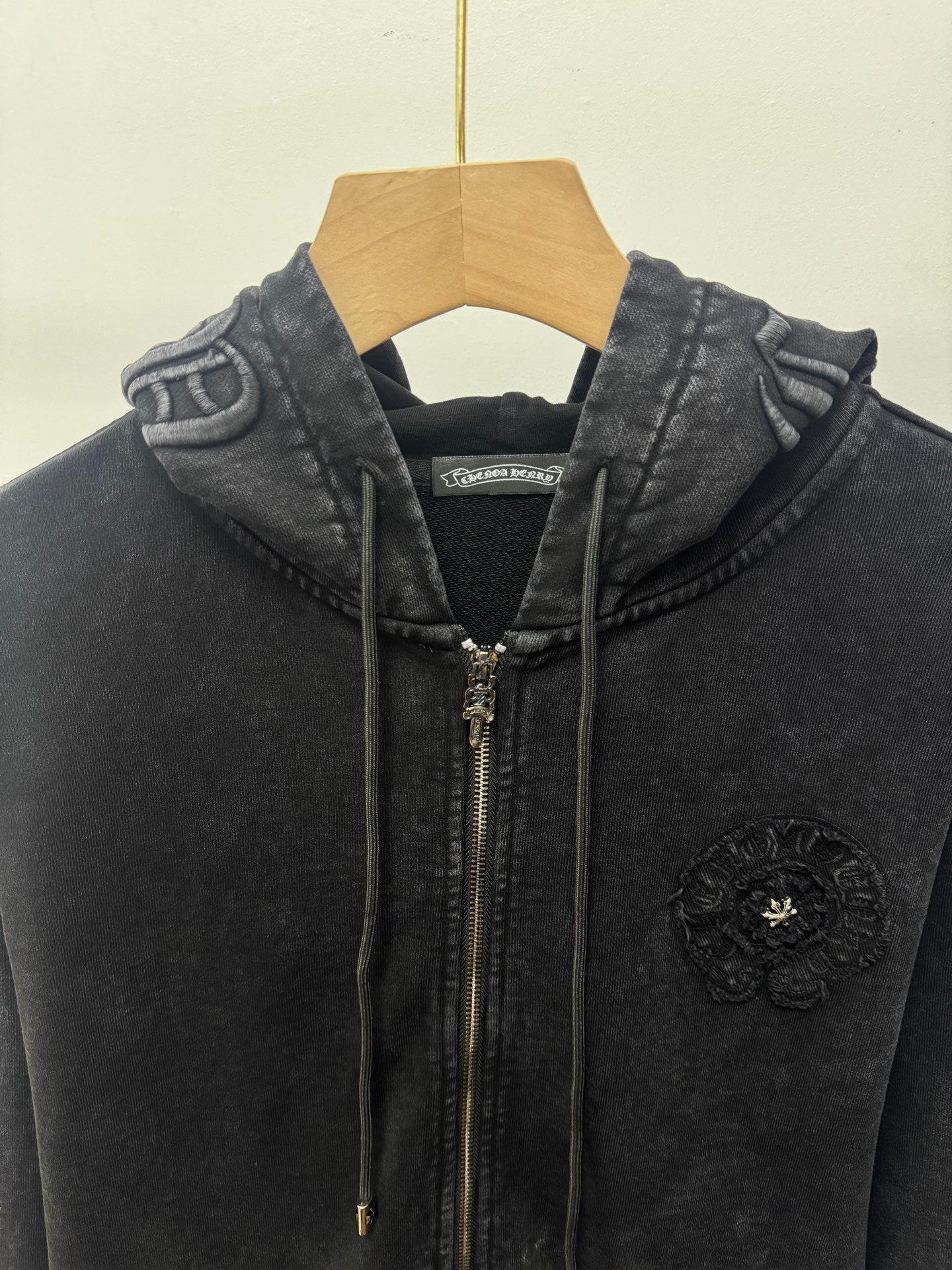Dark zip-up hoodie close-up: hood, front zipper, drawstrings, and chest crest patch.