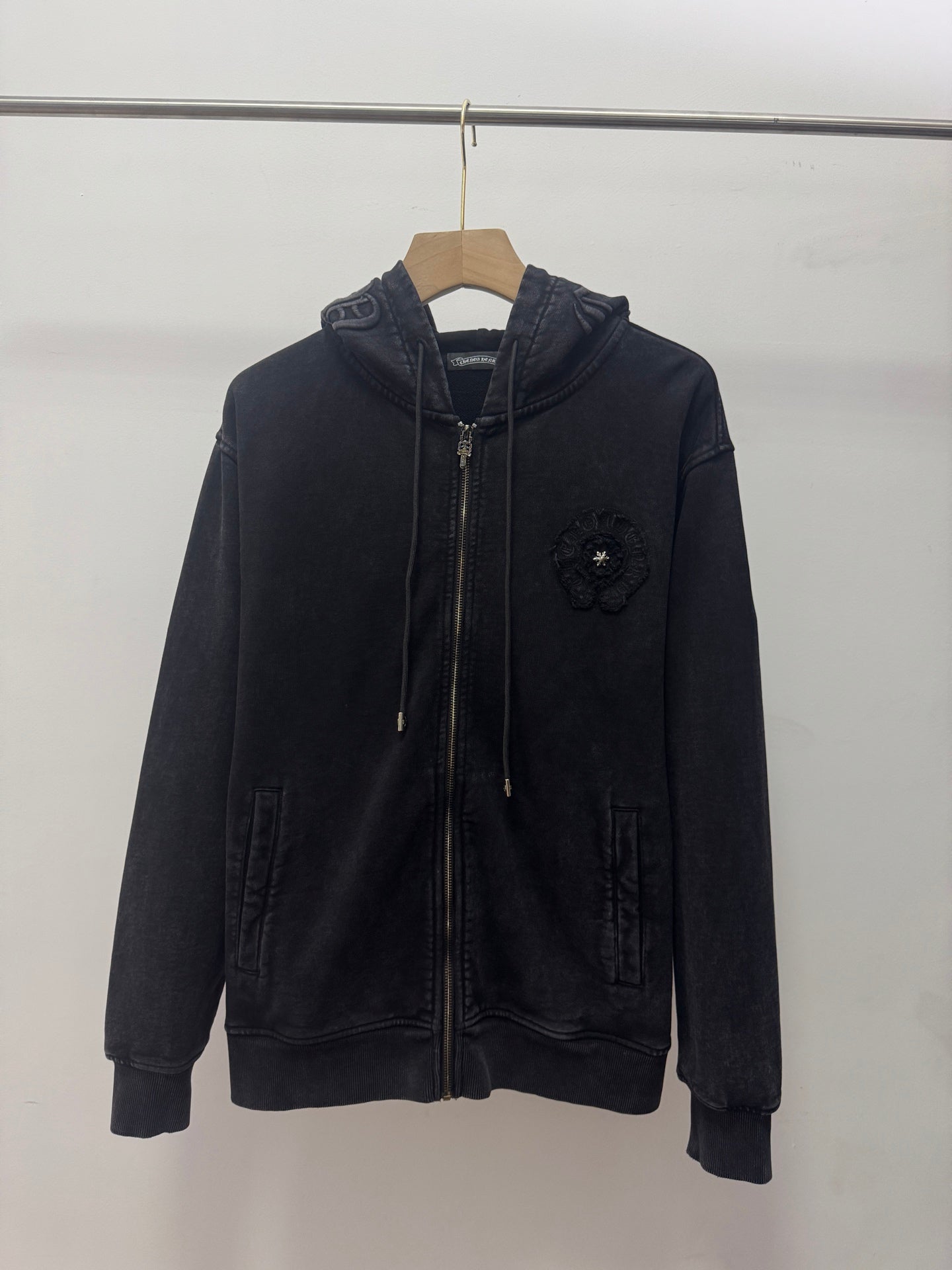 Dark zip-up hoodie with hood, drawstrings, front zipper, and small chest crest patch.