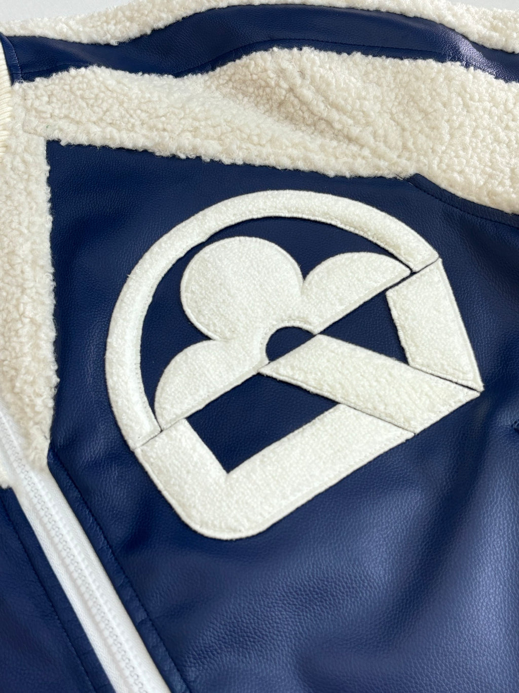 Close-up of cream logo patch on navy faux-leather zip jacket with sherpa trim
