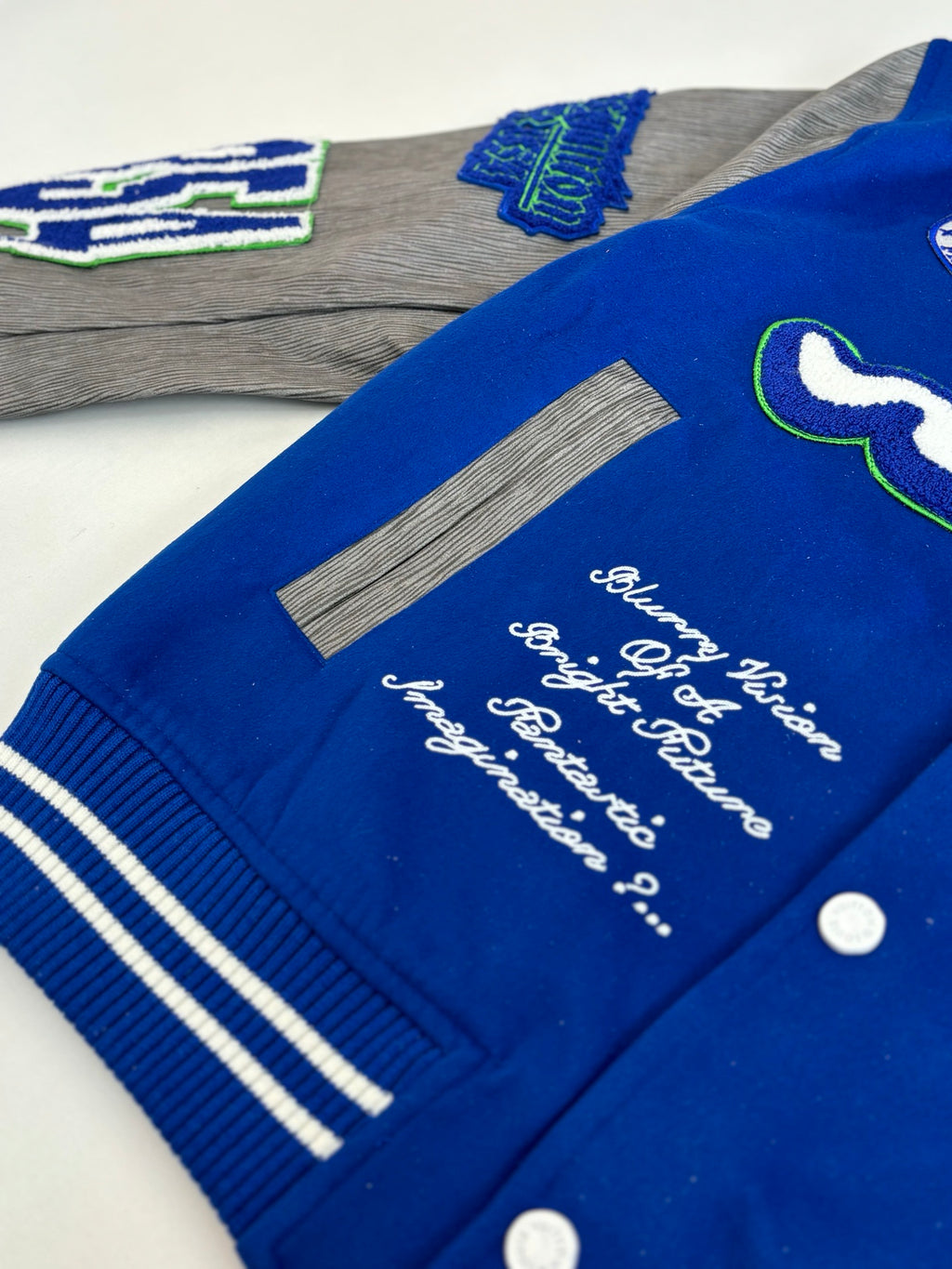 Blue varsity jacket close-up with cursive embroidery, striped ribbing, snap buttons