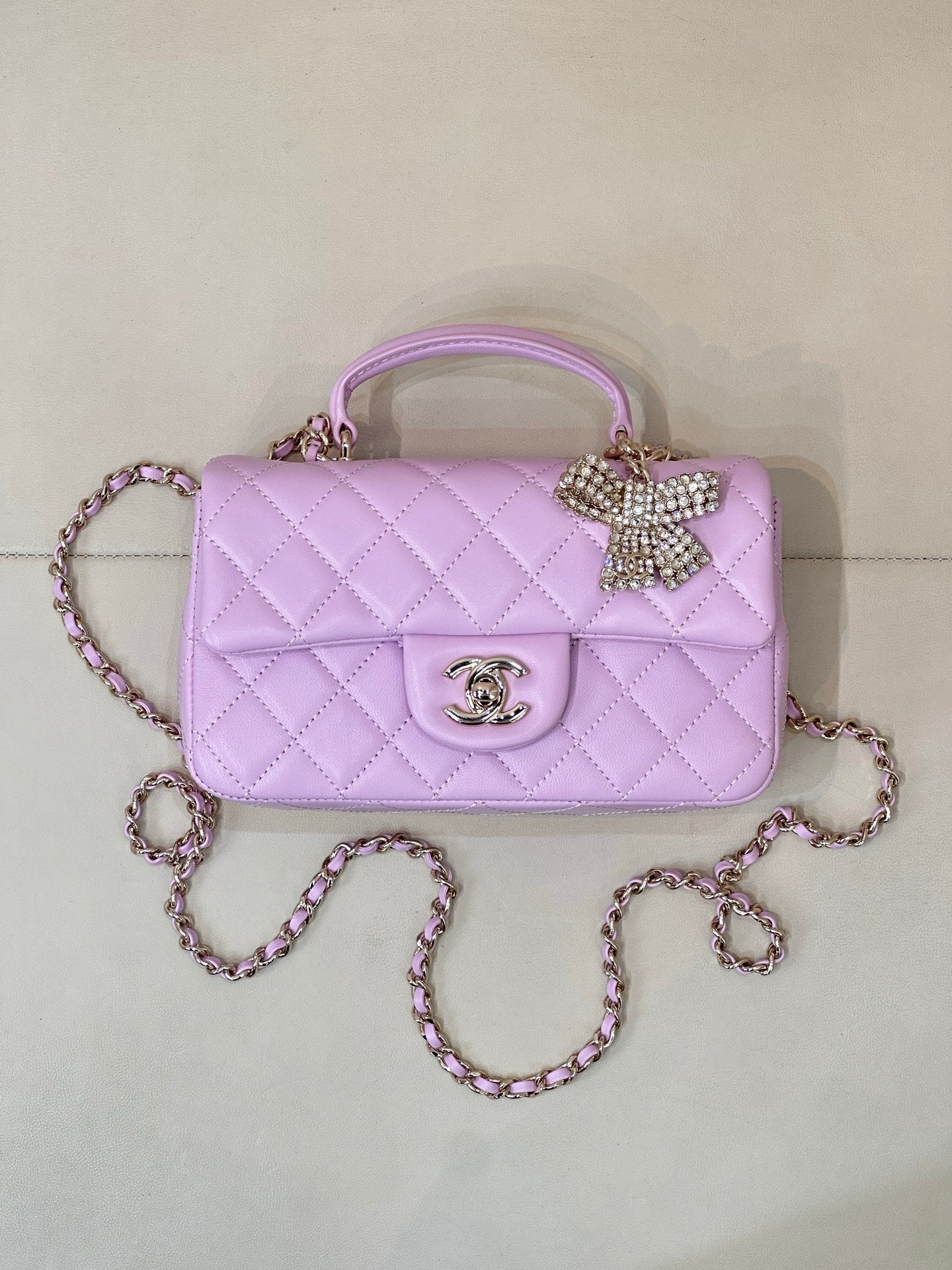Quilted light purple flap handbag with top handle, chain strap, gold logo and bow charm