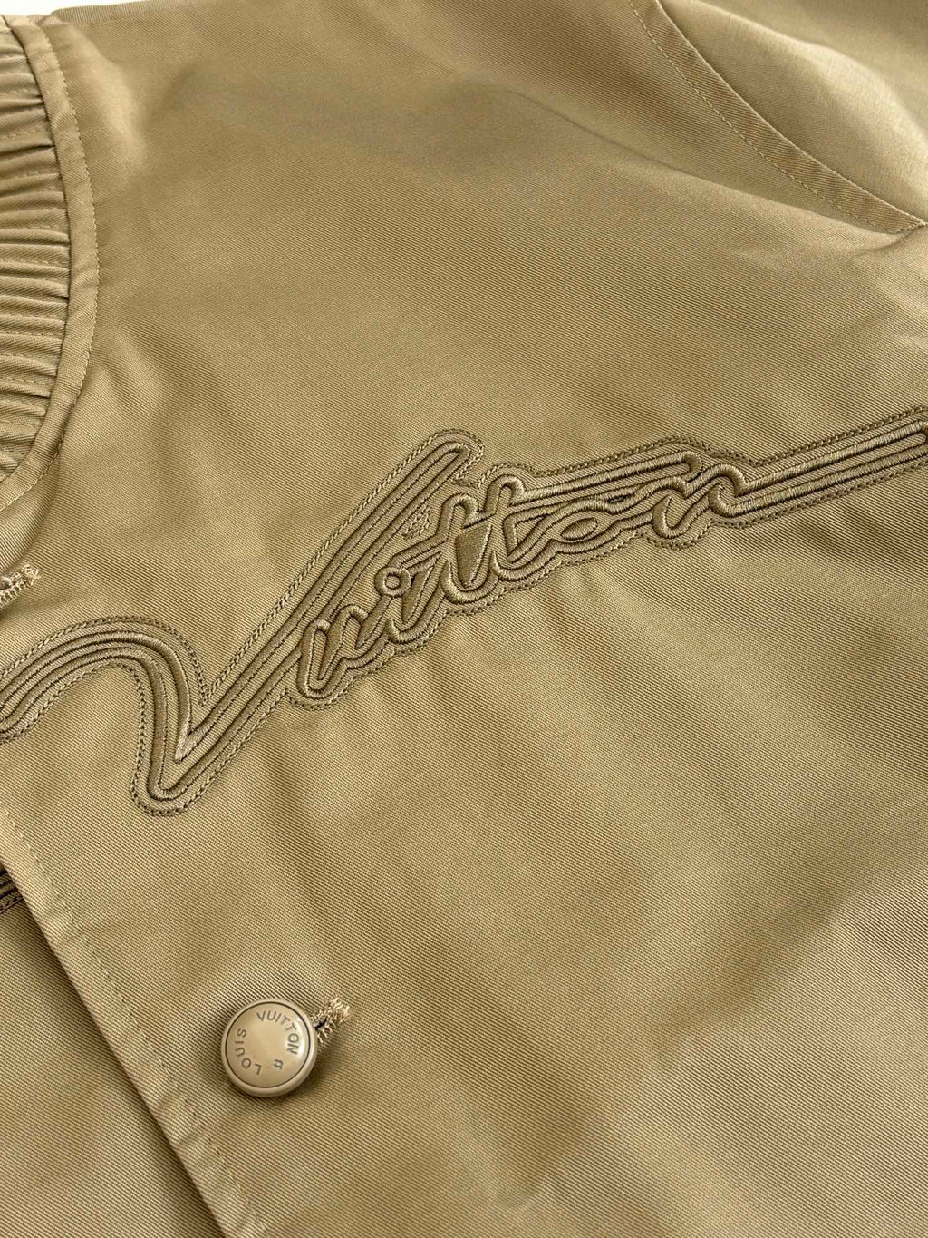 Tan bomber jacket fabric close-up with embroidered trim and round snap button