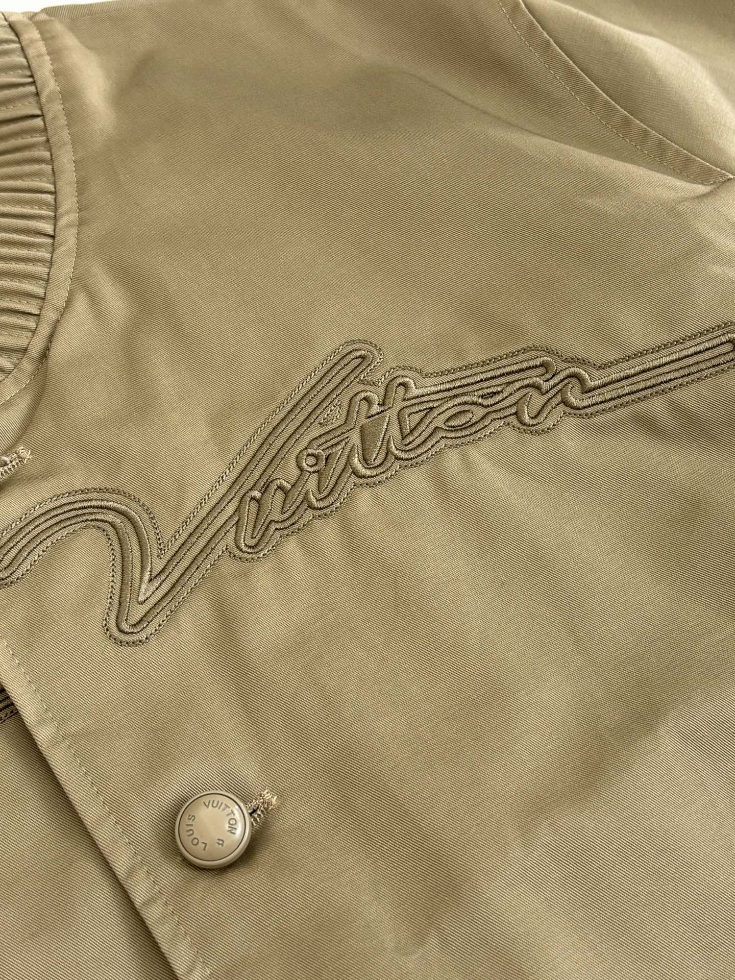 Tan bomber jacket fabric close-up with embroidered trim and round snap button