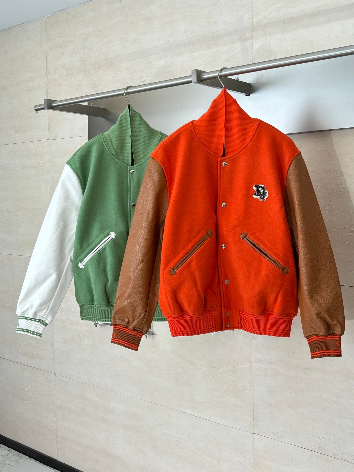 Orange and green varsity jackets on hangers, both with brown leather-look sleeves