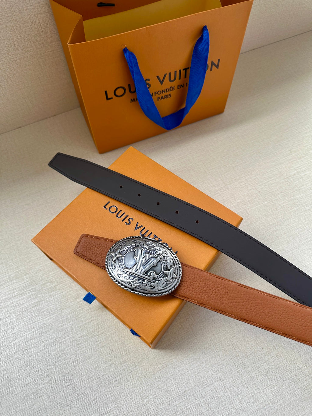 Louis Vuitton belt with tan strap and silver-tone oval logo buckle beside dark belt
