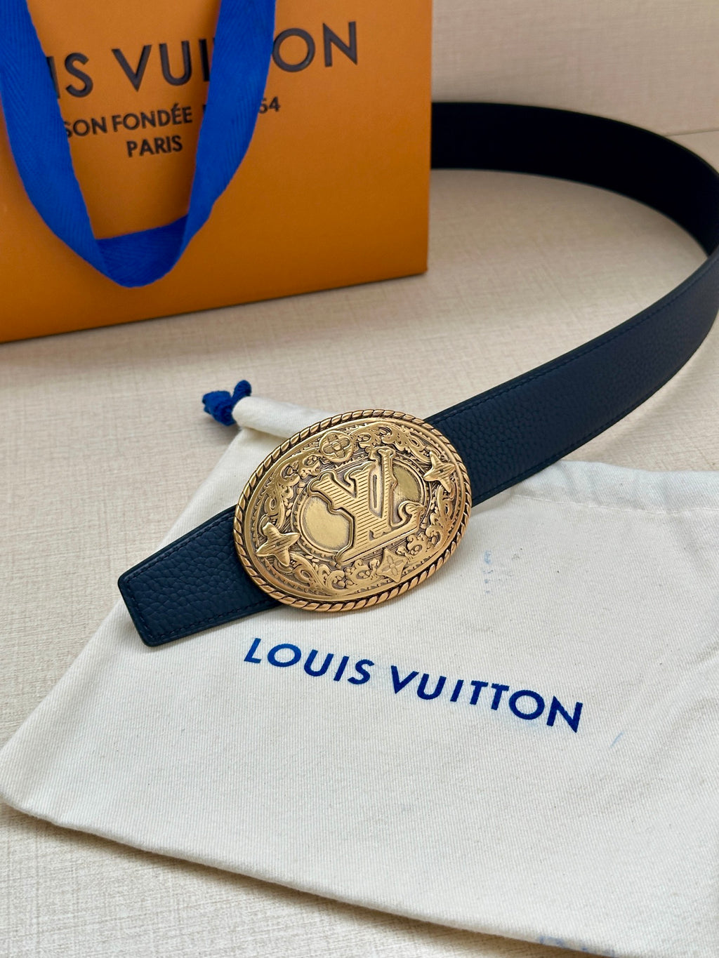 Navy leather belt with gold oval buckle and blue strap ties on dust bag