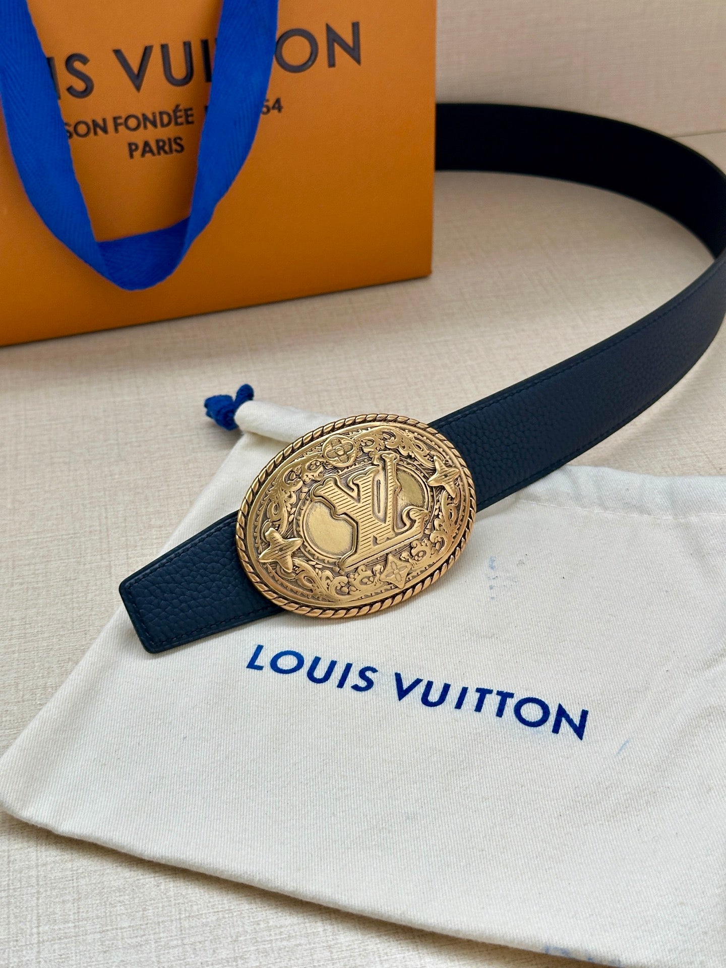 Navy leather belt with gold oval buckle and blue strap ties on dust bag