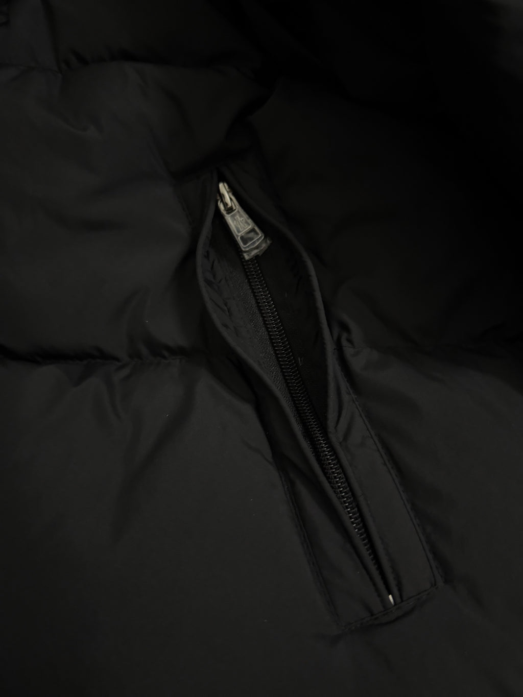 Long puffer coat side pocket zipper with metal pull tab on dark fabric.