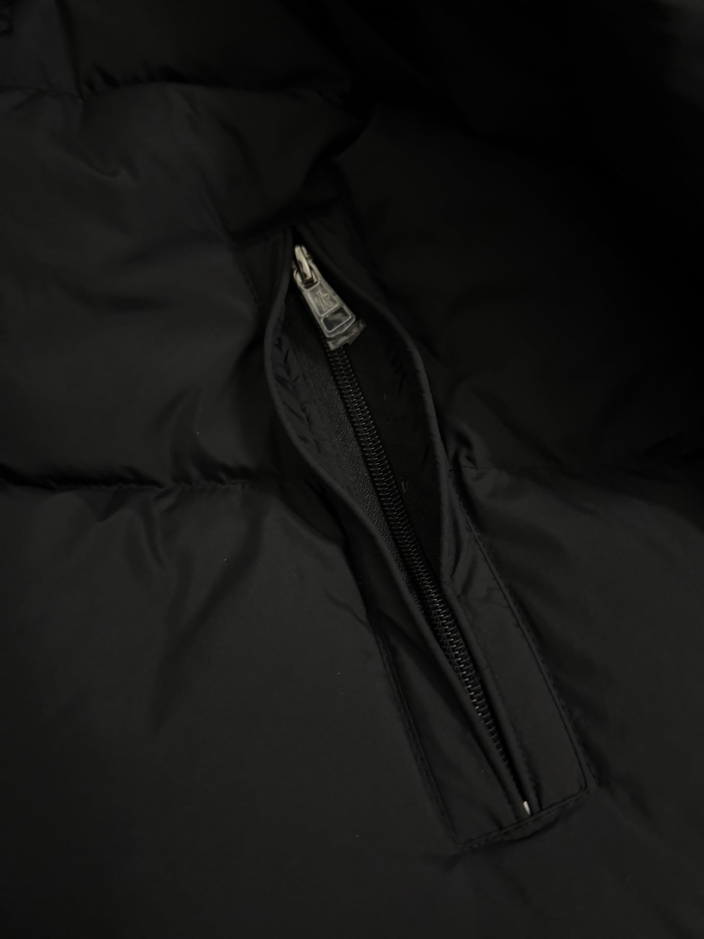 Long puffer coat side pocket zipper with metal pull tab on dark fabric.