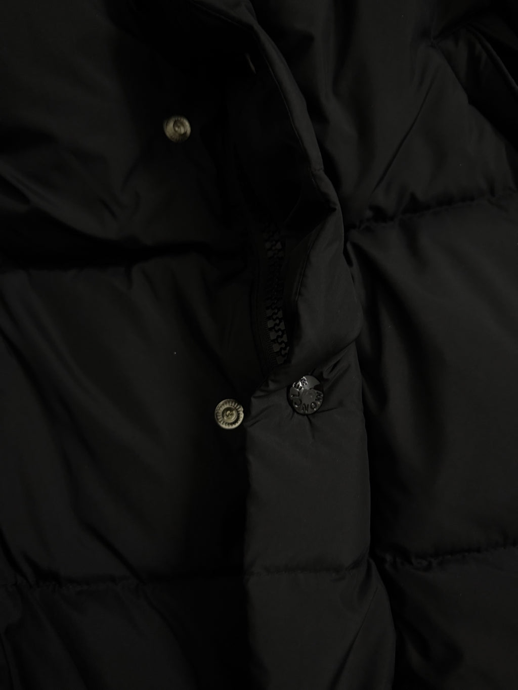 Long puffer coat hood with buttoned placket and metal snaps on dark fabric.