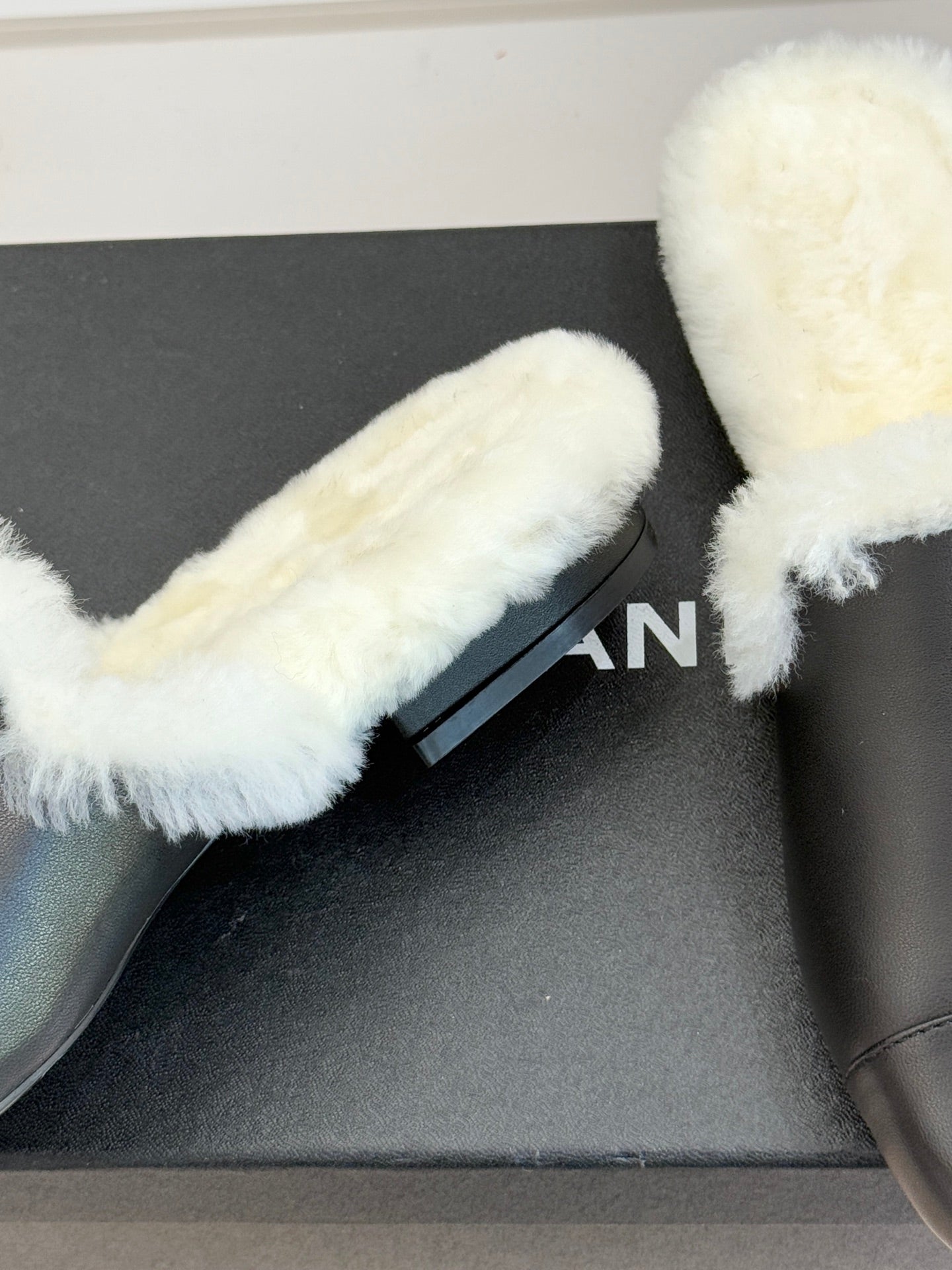 Close-up of black leather mule with white shearling strap and partial logo print