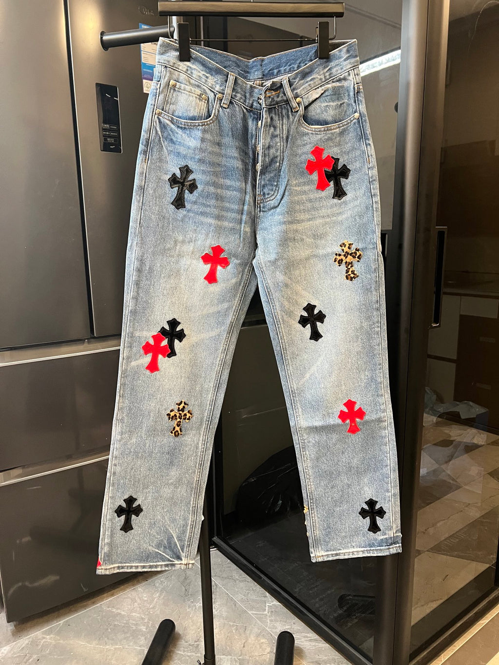 Light-wash denim pants with red and black cross patches, front view on hanger