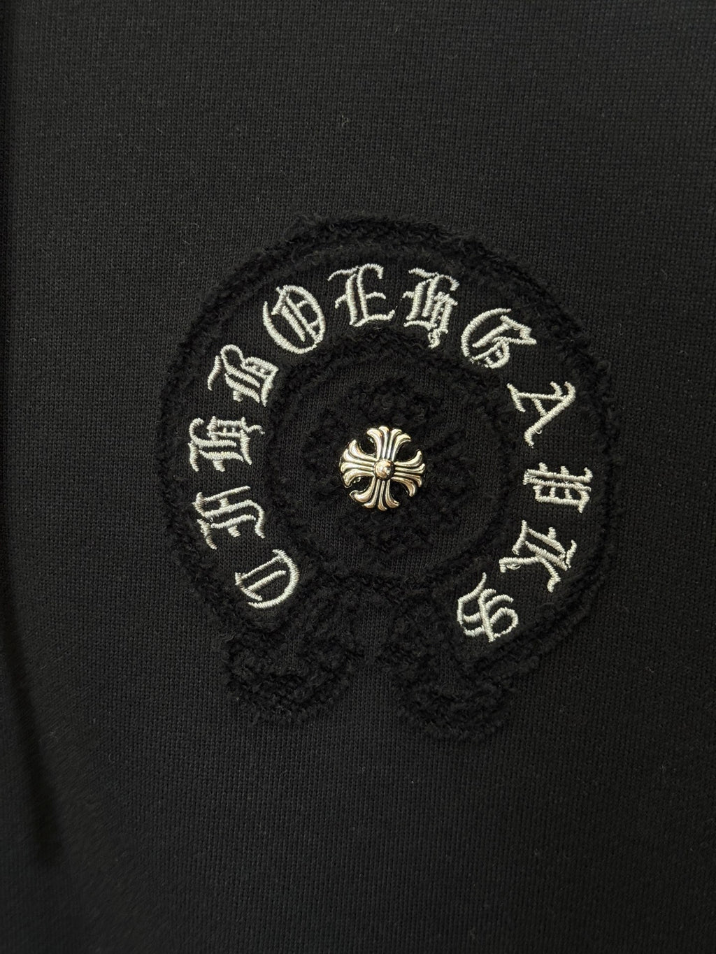Embroidered circular patch on black hoodie, gold-toned symbols and small center flower.