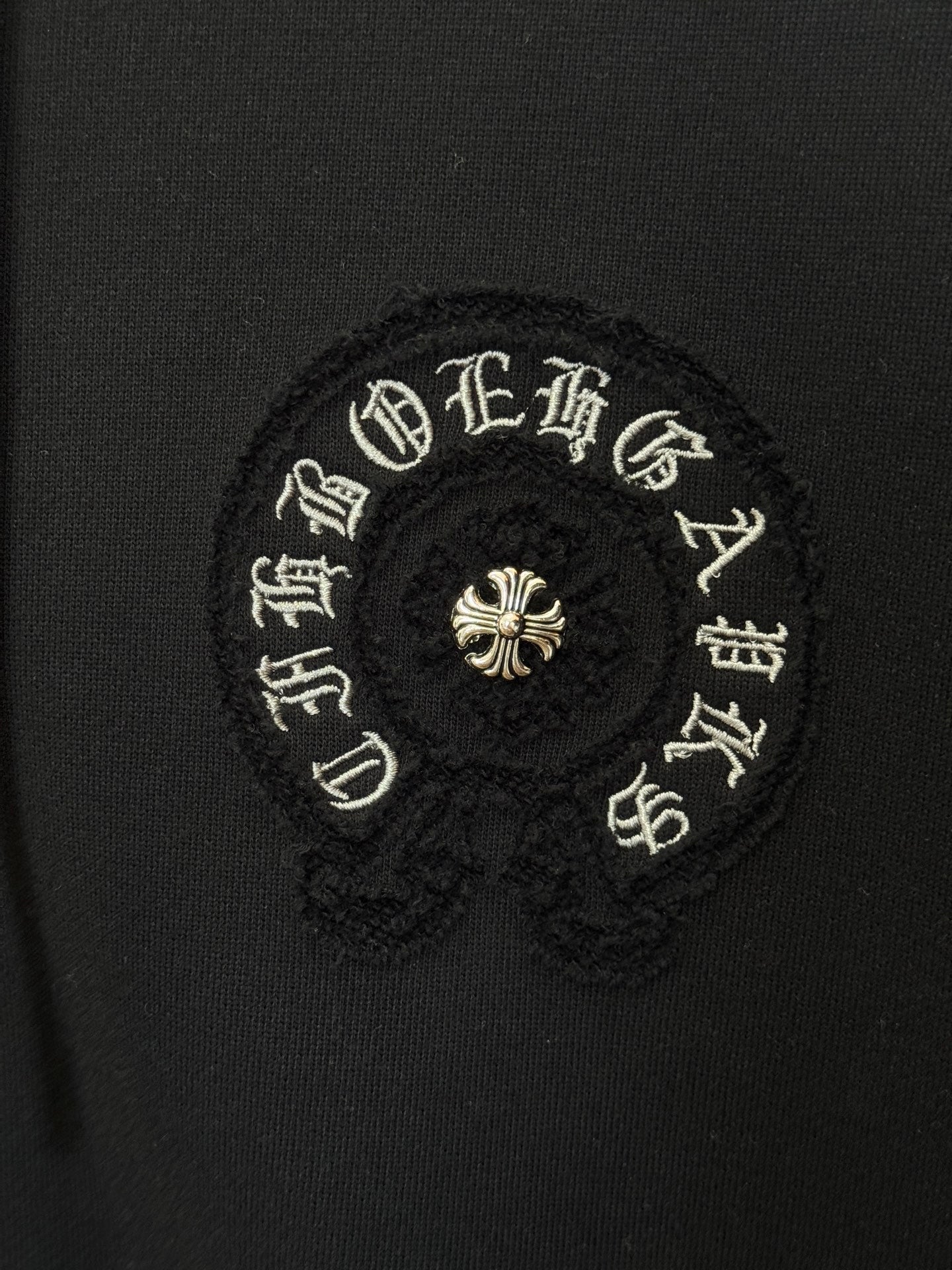 Embroidered circular patch on black hoodie, gold-toned symbols and small center flower.