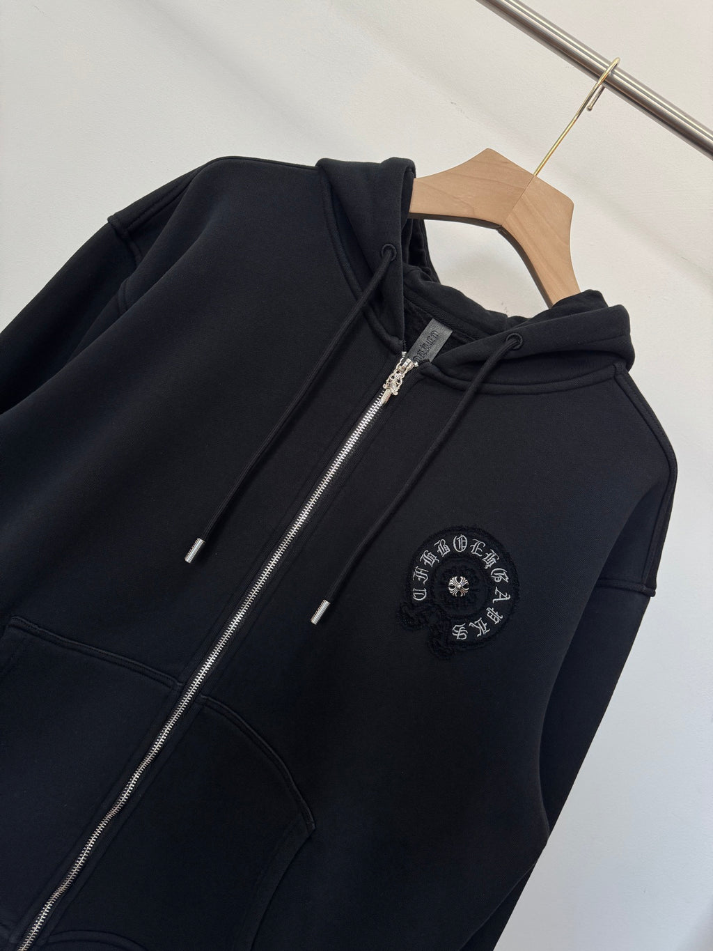 Black zip-up hoodie detail view with metal zipper pull and circular chest emblem.