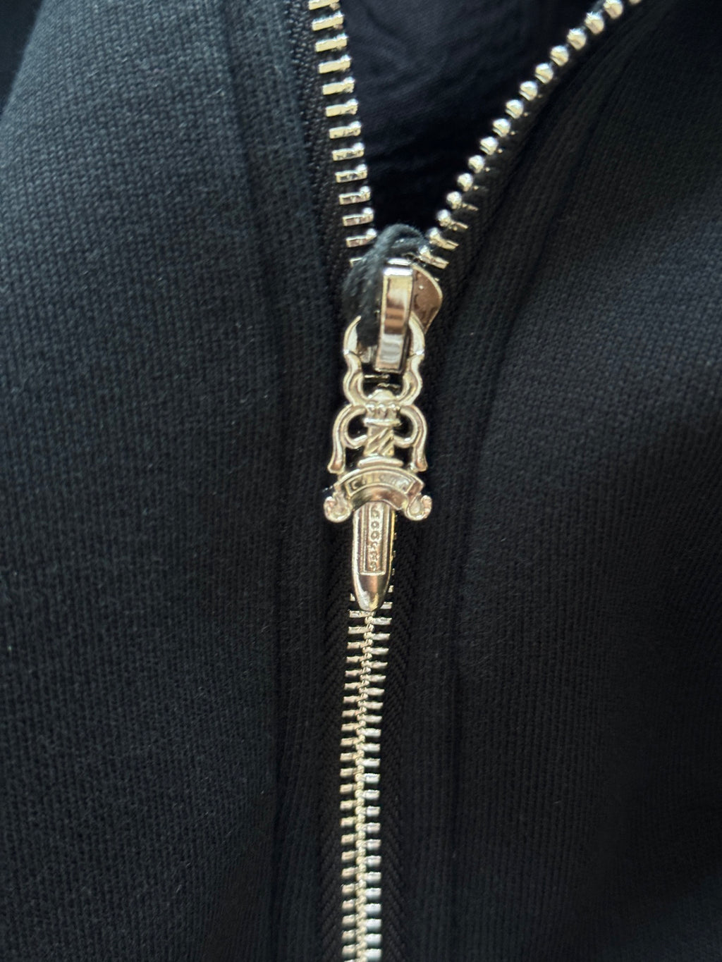 Close-up of metal zipper pull on black hoodie with stitched teeth and keychain-style charm.