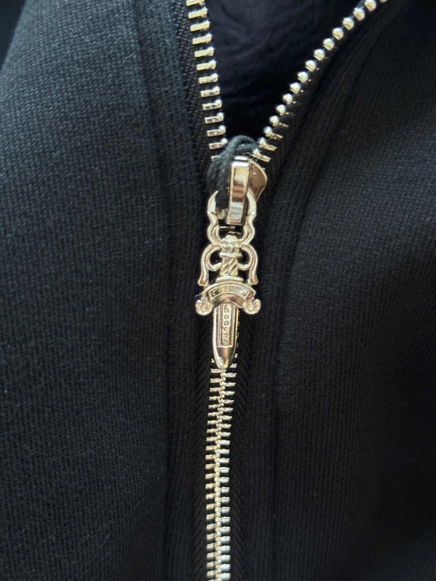 Close-up of metal zipper pull on black hoodie with stitched teeth and keychain-style charm.
