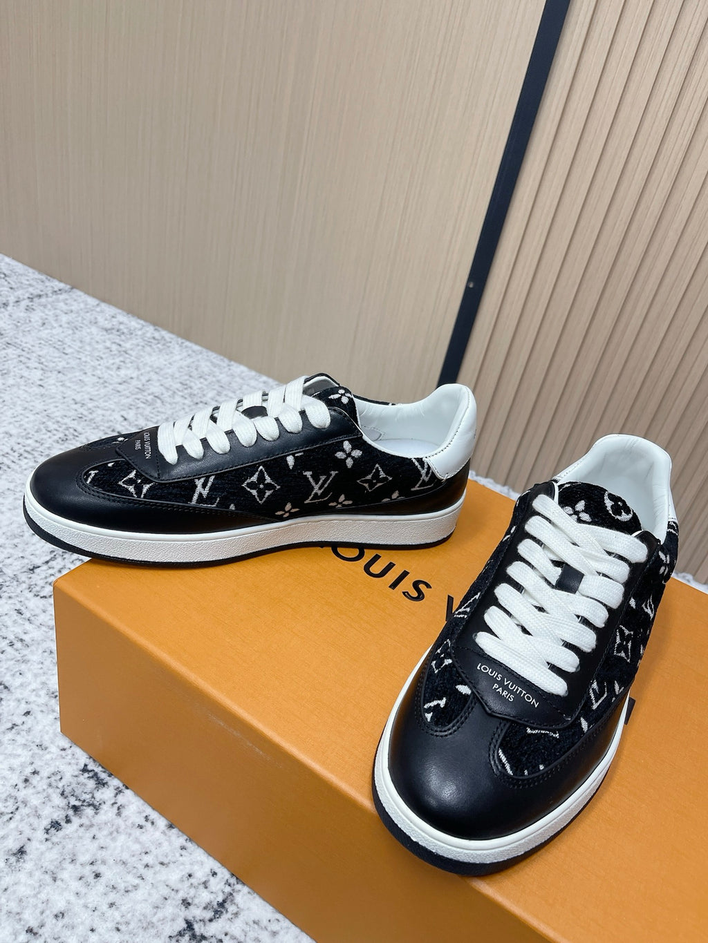 Black low-top sneakers with white laces and monogram-style pattern, shown on shoe box.