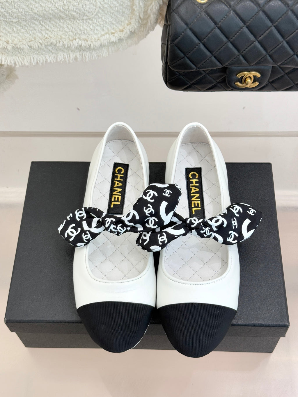 White Chanel ballet flats with black bow featuring CHANEL print on top.
