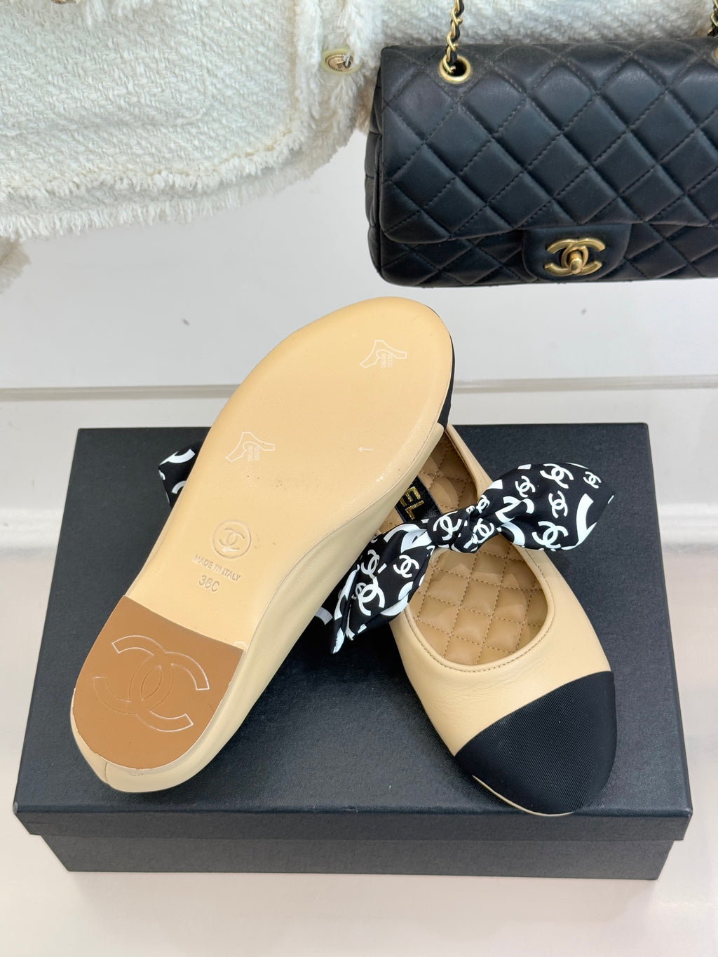Beige flat with navy toe cap and black logo bow; outsole view on display box