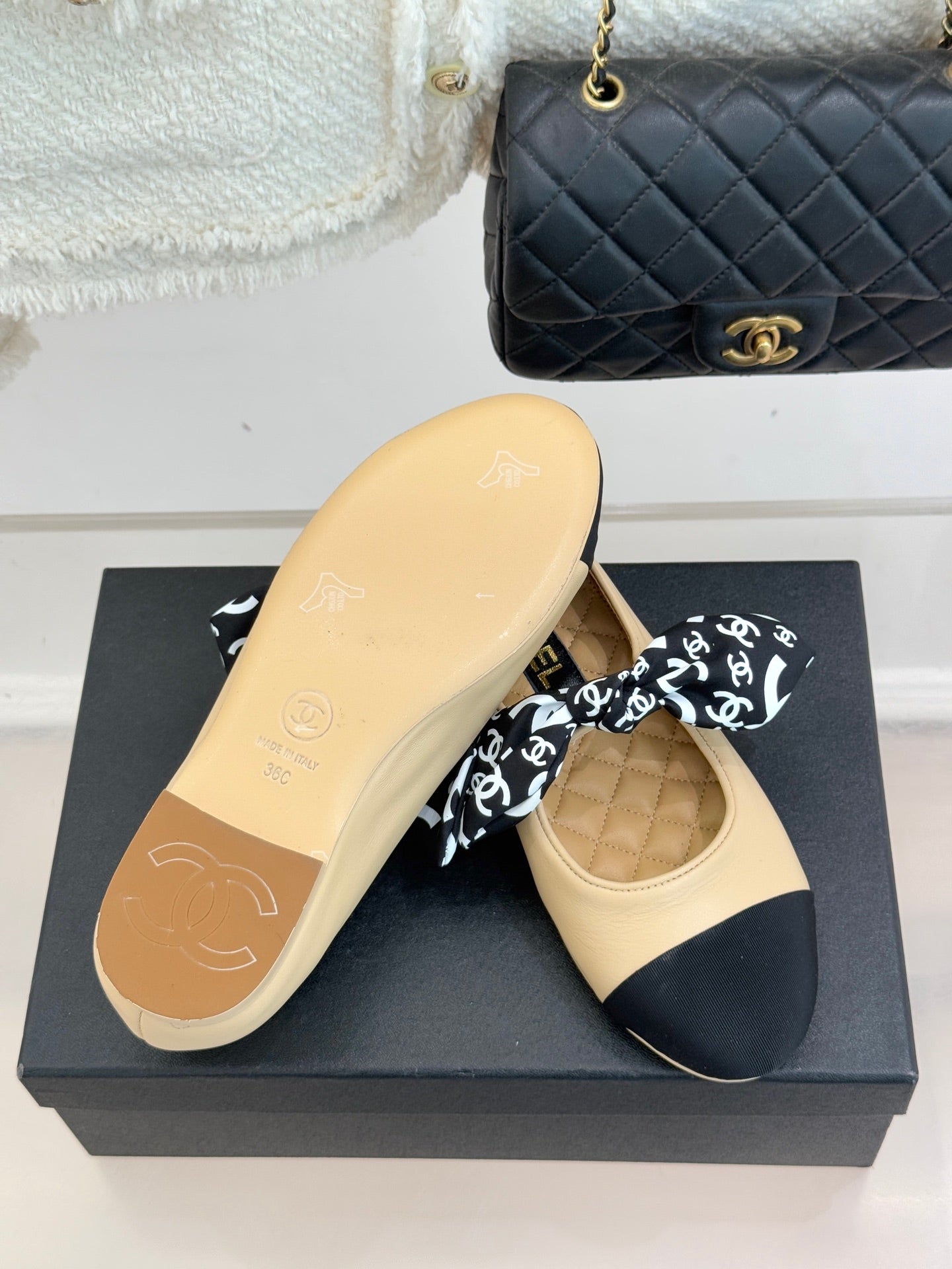 Beige flat with navy toe cap and black logo bow; outsole view on display box