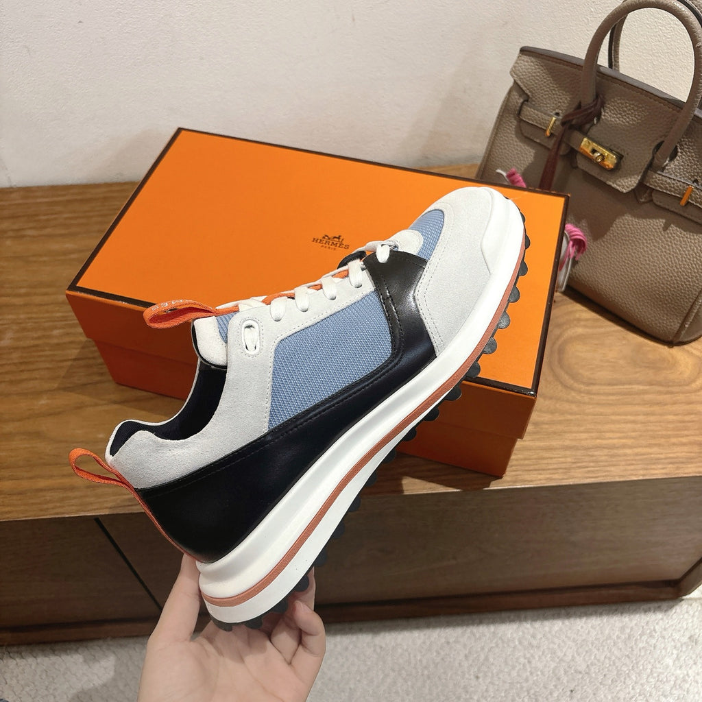 Unisex sneaker held by hand over shoebox; white, blue, black color-block with orange laces