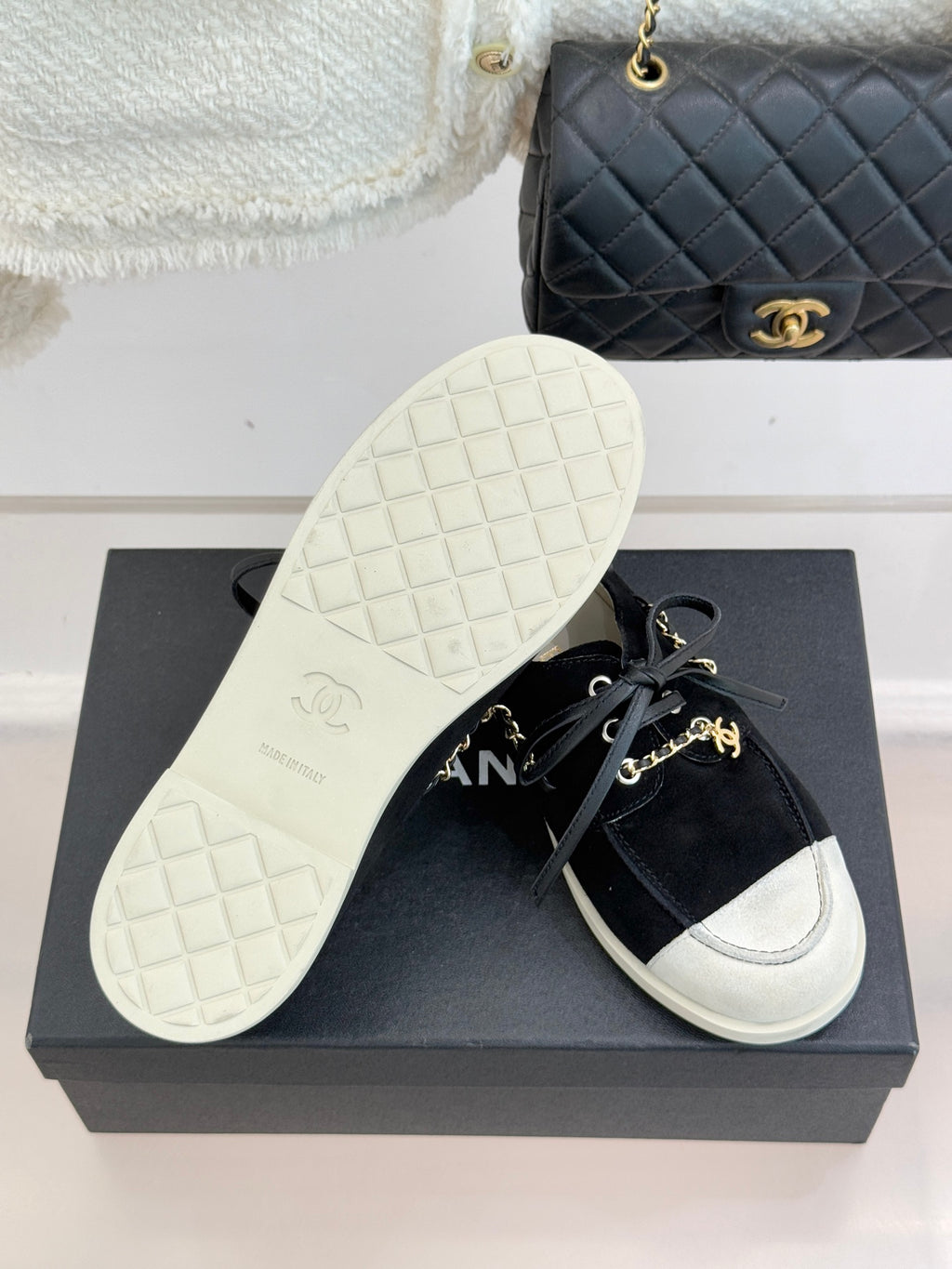 Black suede sneakers on a box, with white treaded outsole and gold chain trim