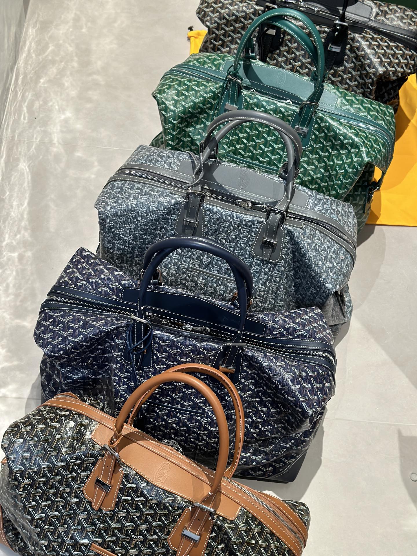 Goyard duffel bags in green, navy, and gray stacked with top handles and straps