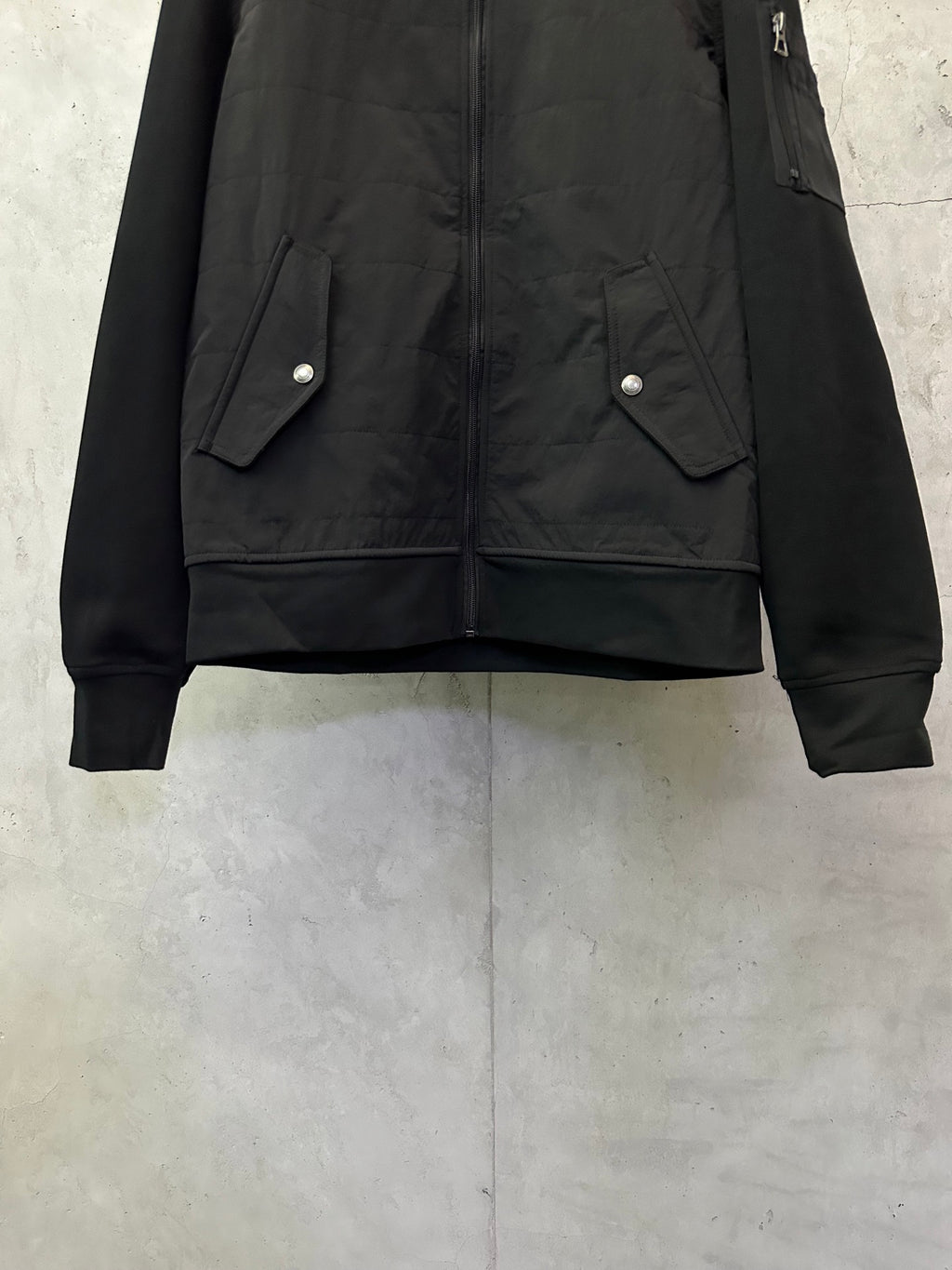 Black bomber jacket with snap-flap pockets and ribbed cuffs, front lower view.