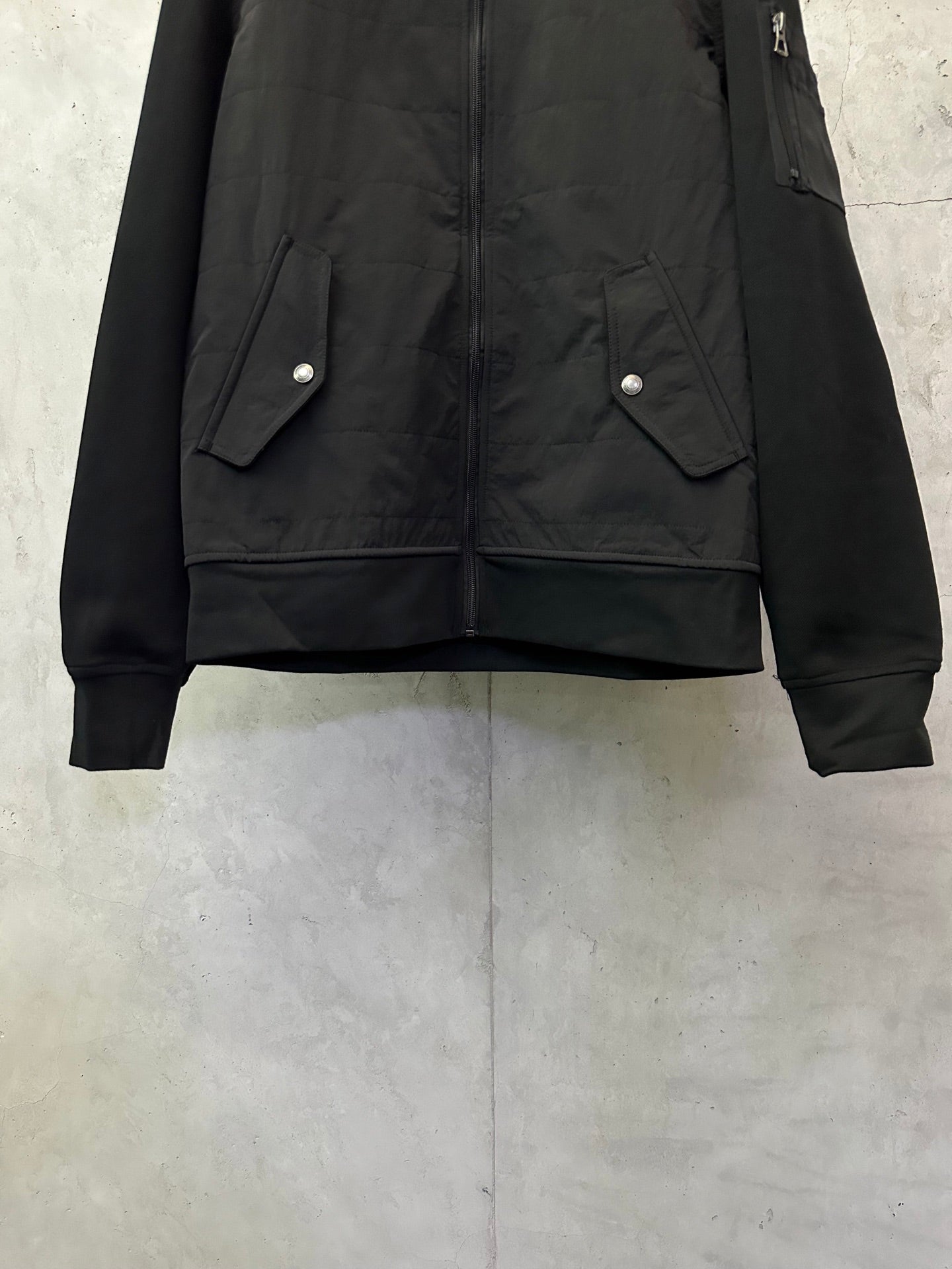 Black bomber jacket with snap-flap pockets and ribbed cuffs, front lower view.