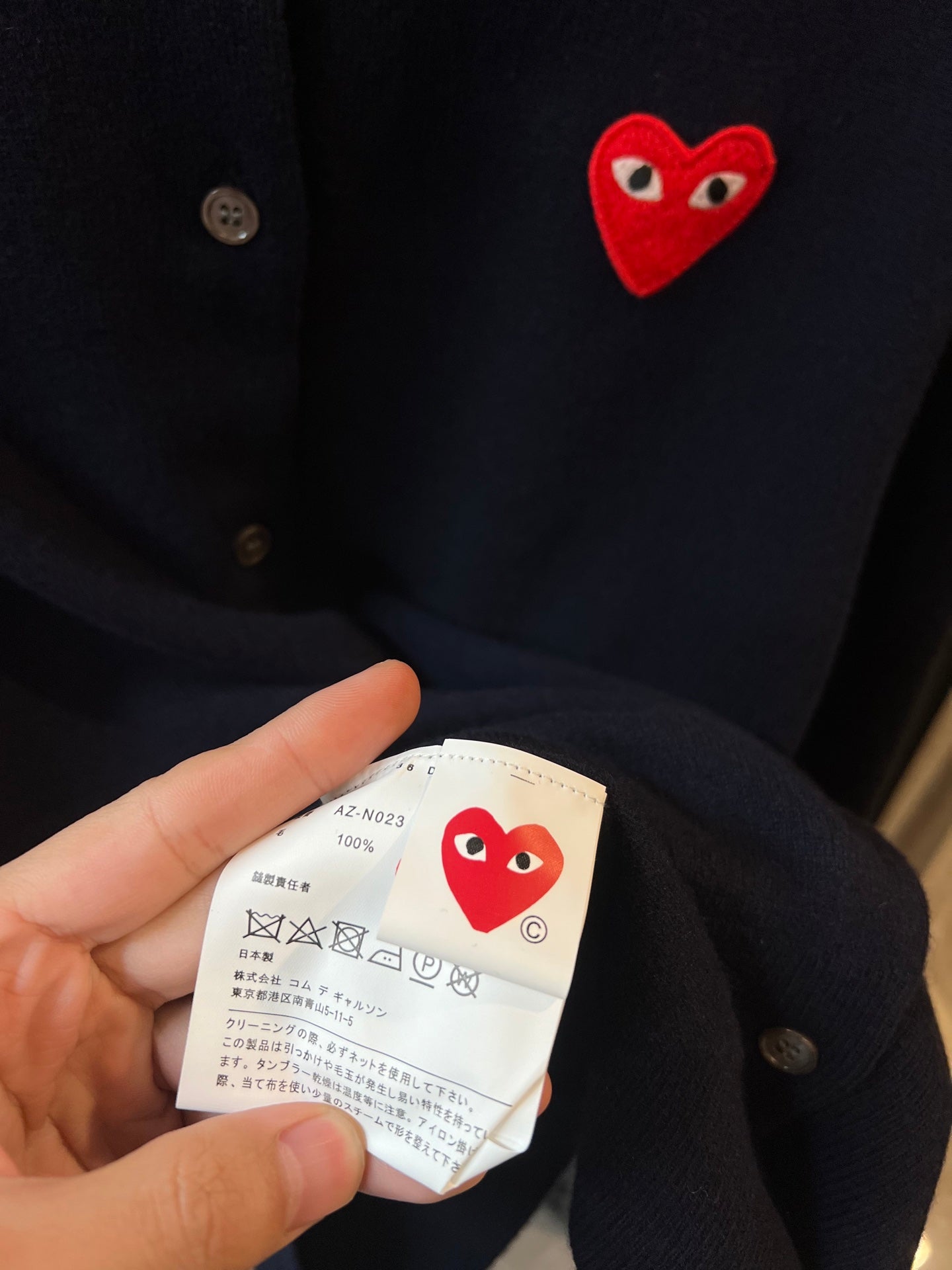 Dark navy knit cardigan with red heart patch and visible care label held in hand
