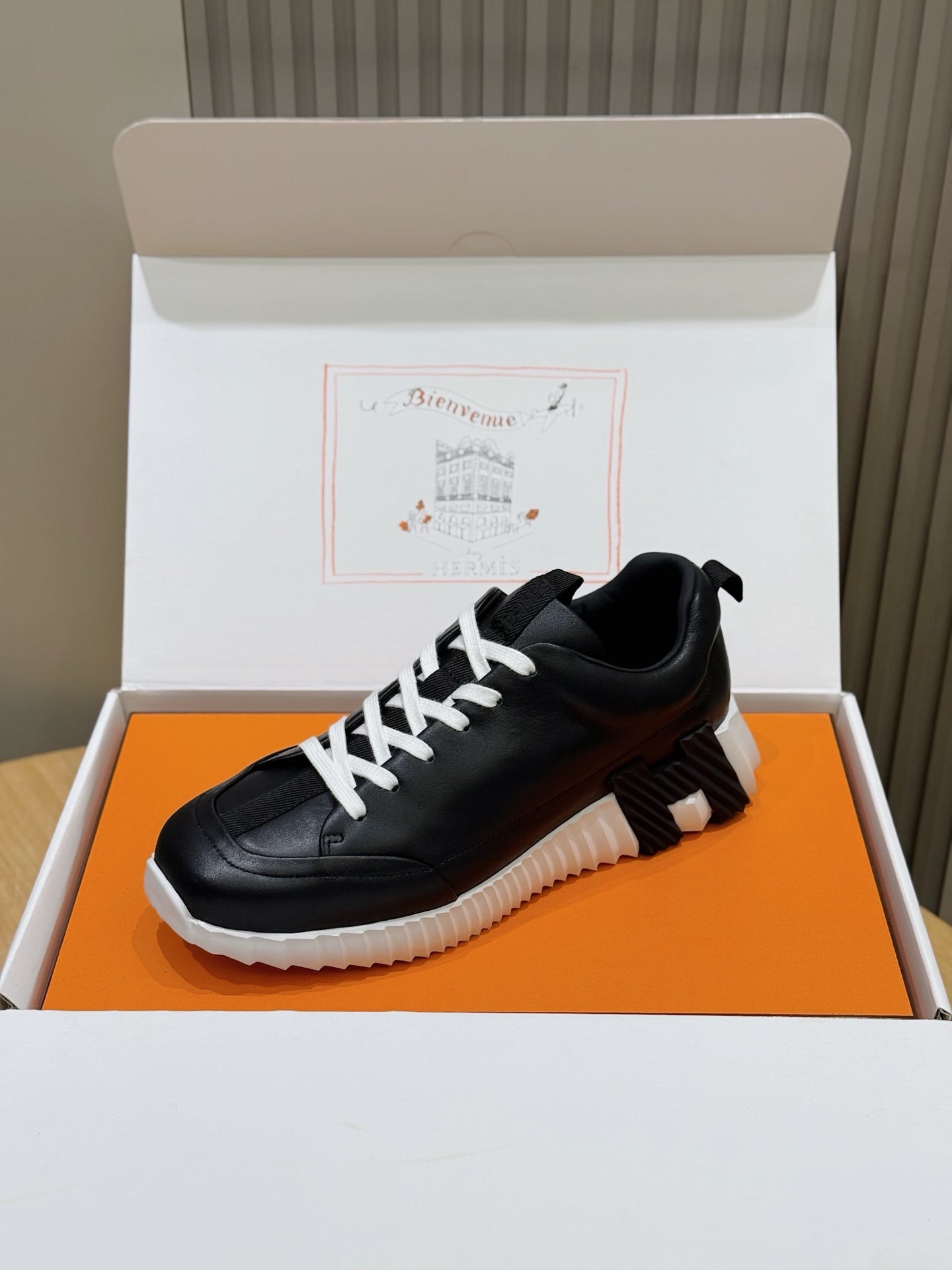Black lace-up sneaker with white laces, shown in orange shoe box