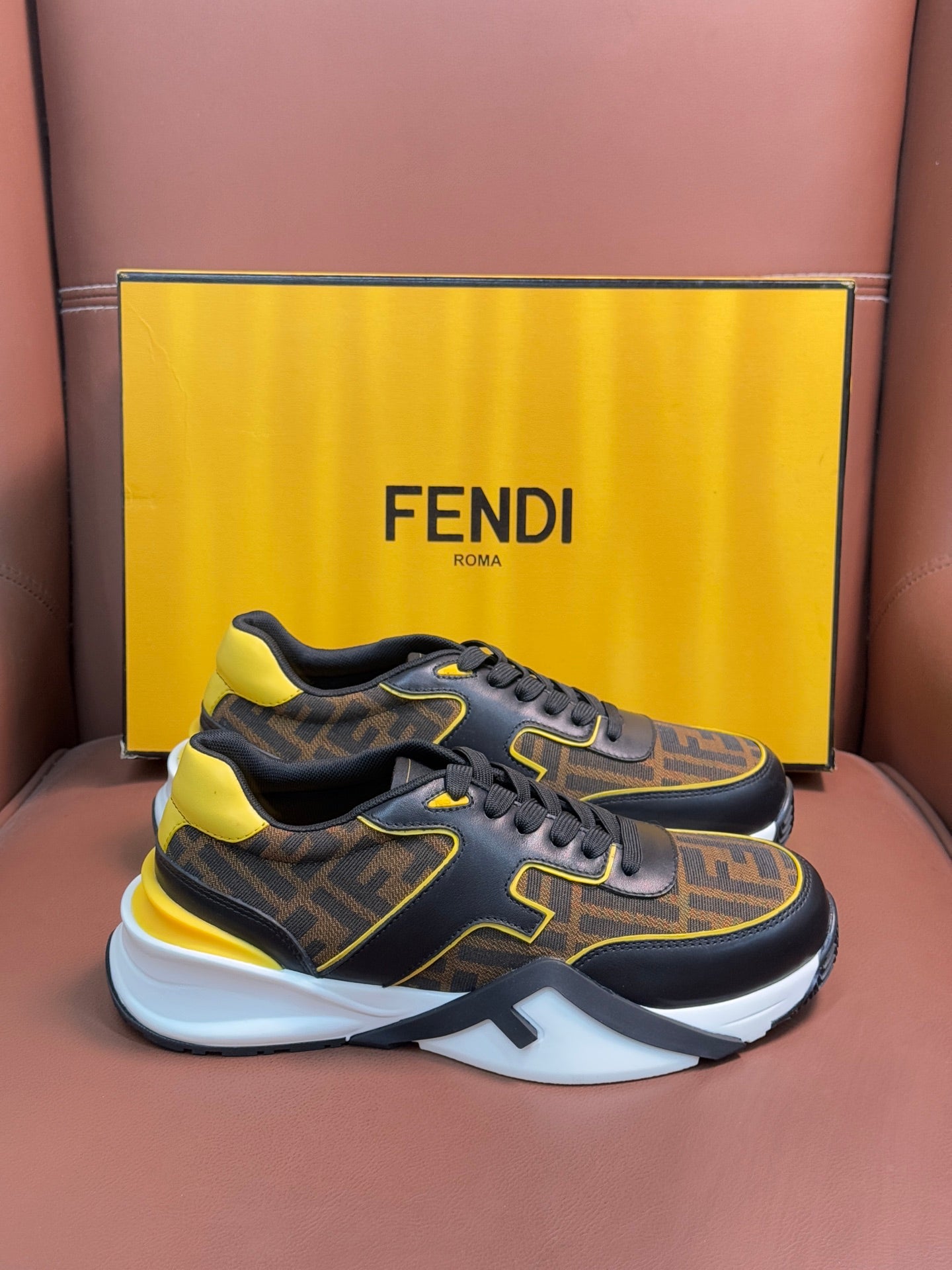 Fendi unisex sneakers on box, dark FF fabric with yellow details and thick white outsole.