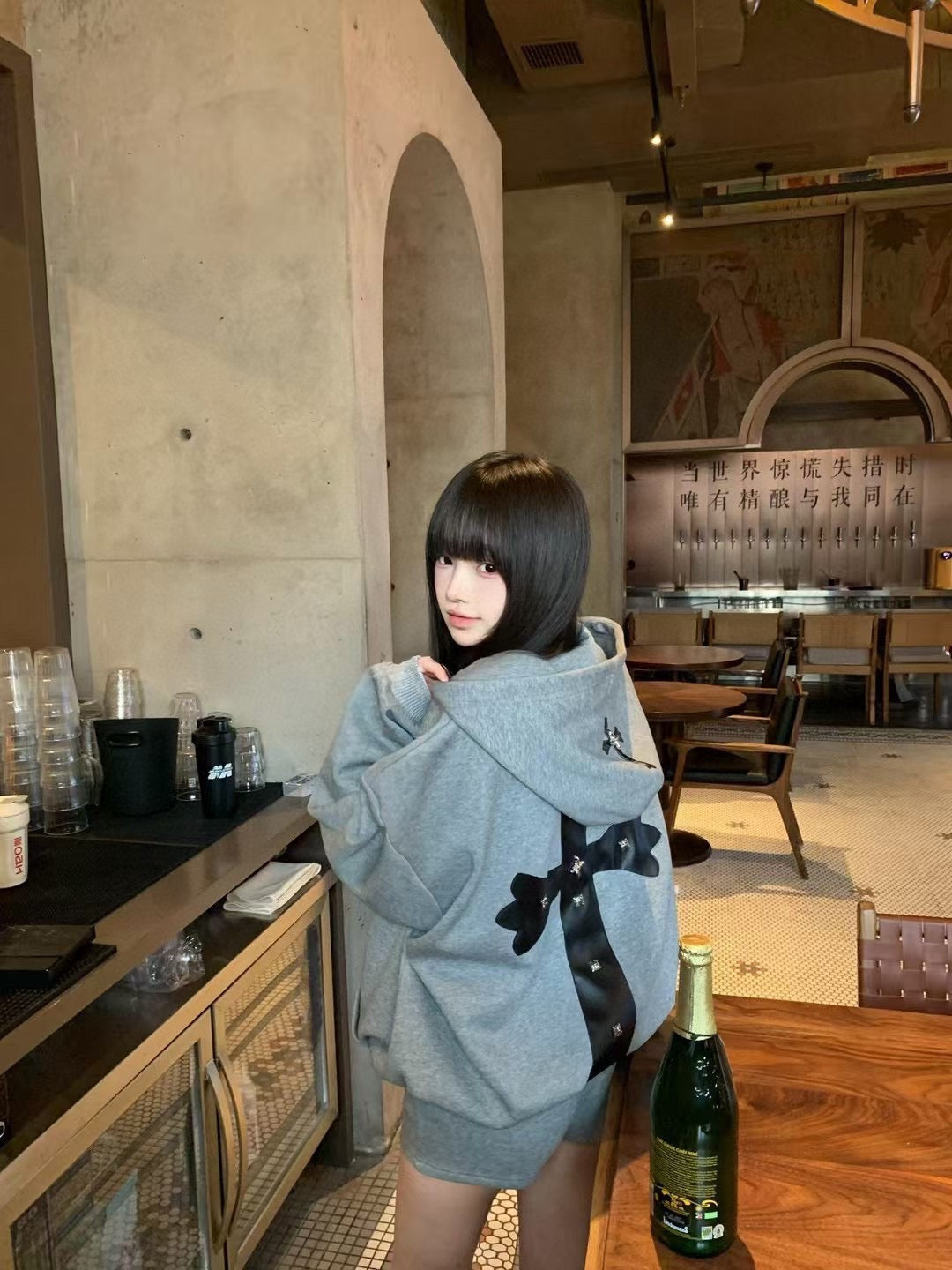 Gray hoodie with black bow-tie accents, hood up on model indoors