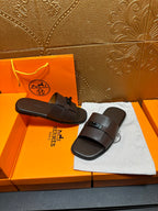 Pair of dark slide sandals with buckle strap, resting on white dust bag.