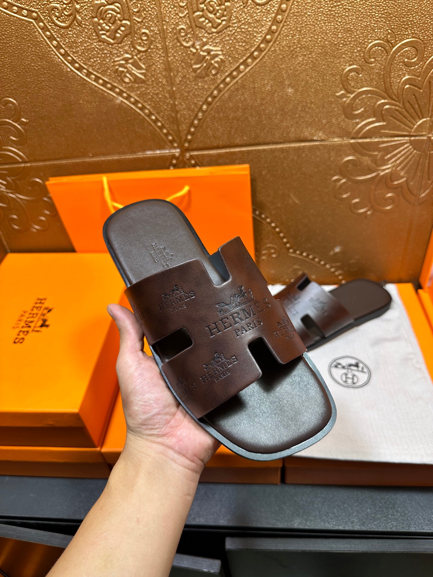 Brown leather slide sandal held in hand, embossed logo on upper strap
