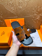 Hermes slide sandal close-up held in hand, tan embossed straps on dark sole.